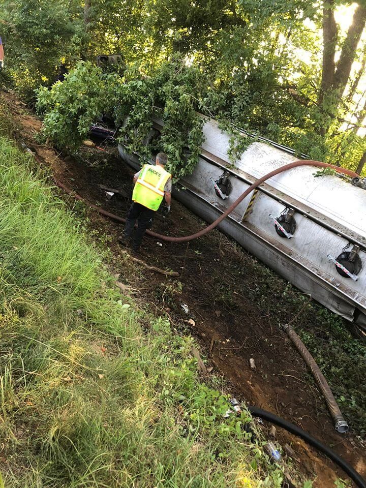 A man is standing next to a tanker truck that has fallen off the road.