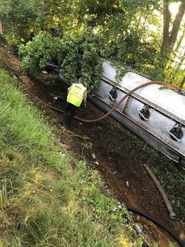 A man is standing next to a tanker truck that has fallen off the road.