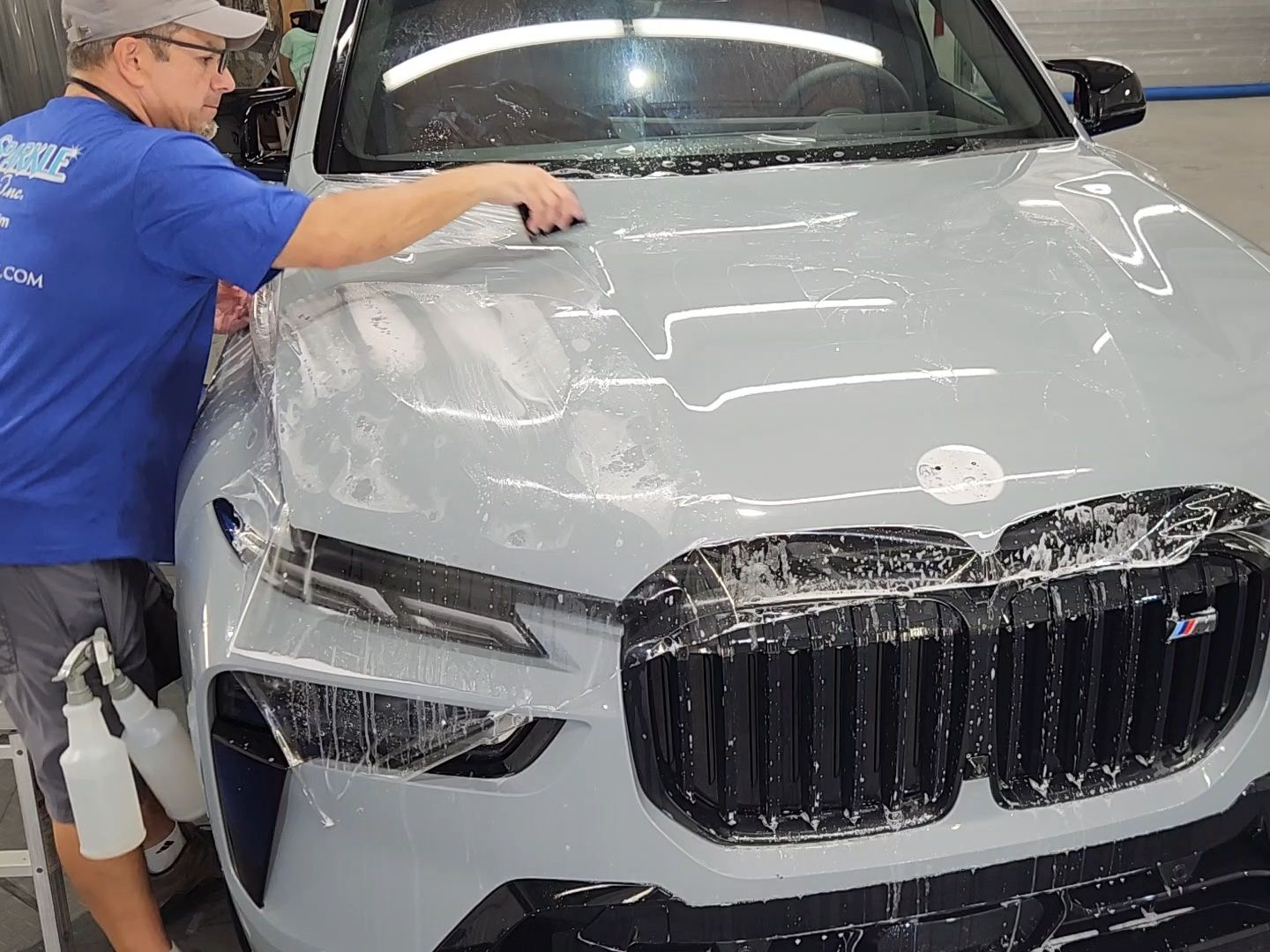 Man applying protective film to the hood of a light gray BMW SUV; spraying water, in a shop.