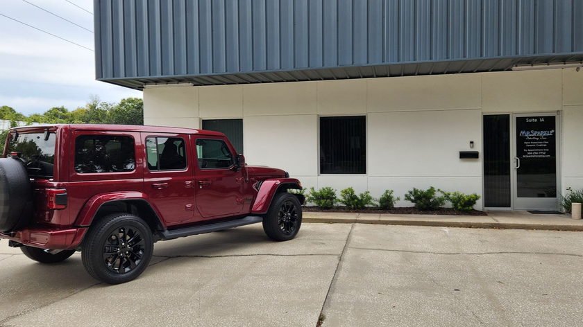 Red Jeep parked in front of a white and gray building with a door and small windows.