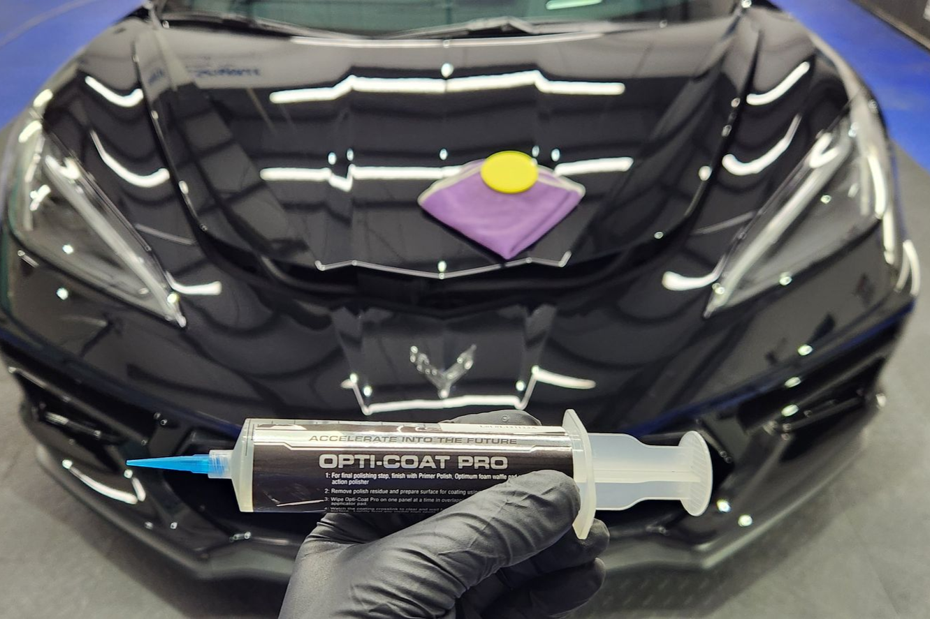 Gloved hand holding Opti-Coat Pro syringe over a shiny black car hood with an applicator pad.