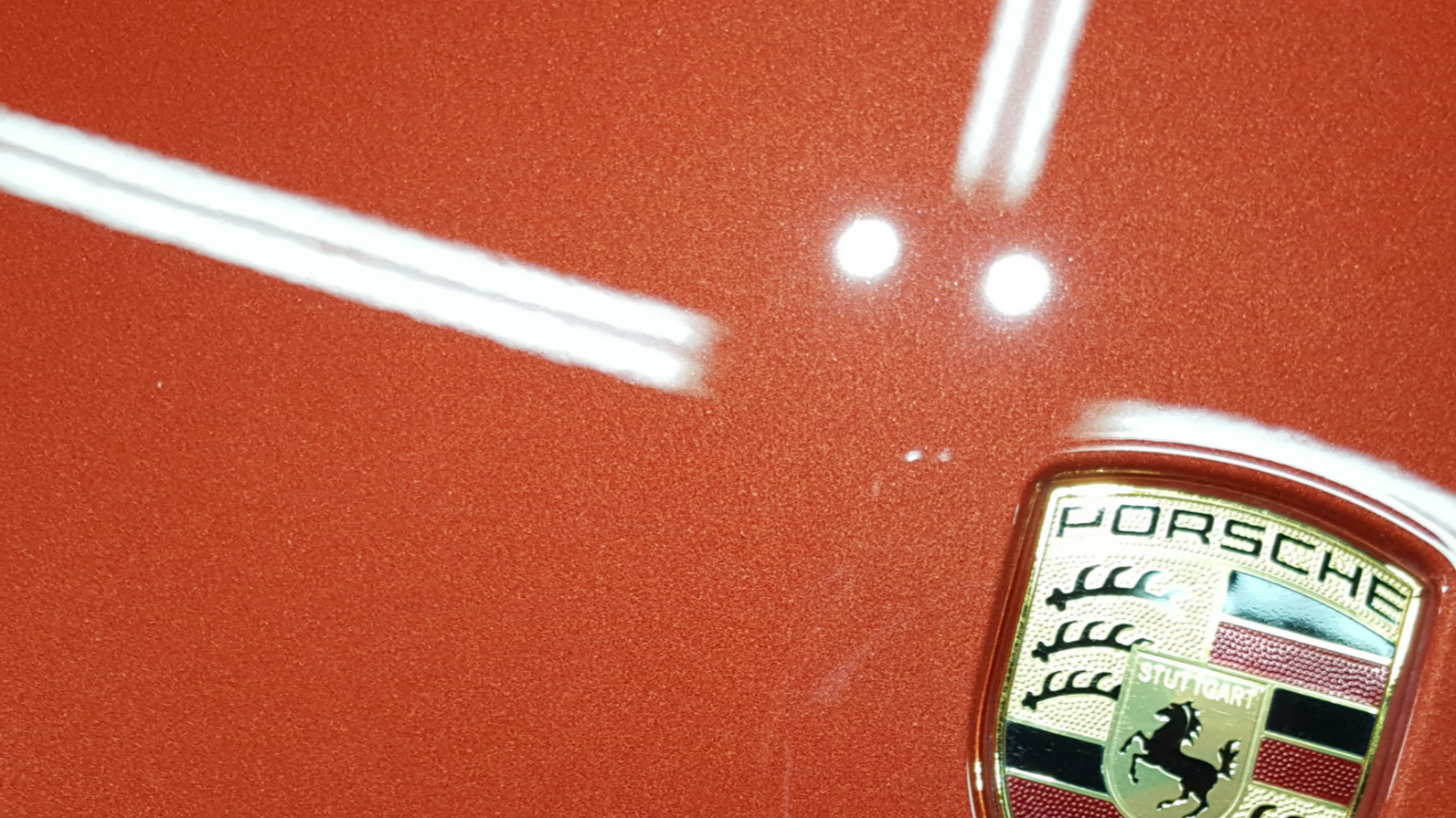 Close-up of orange Porsche car body with the brand's emblem. White reflective lines are seen on the paint.