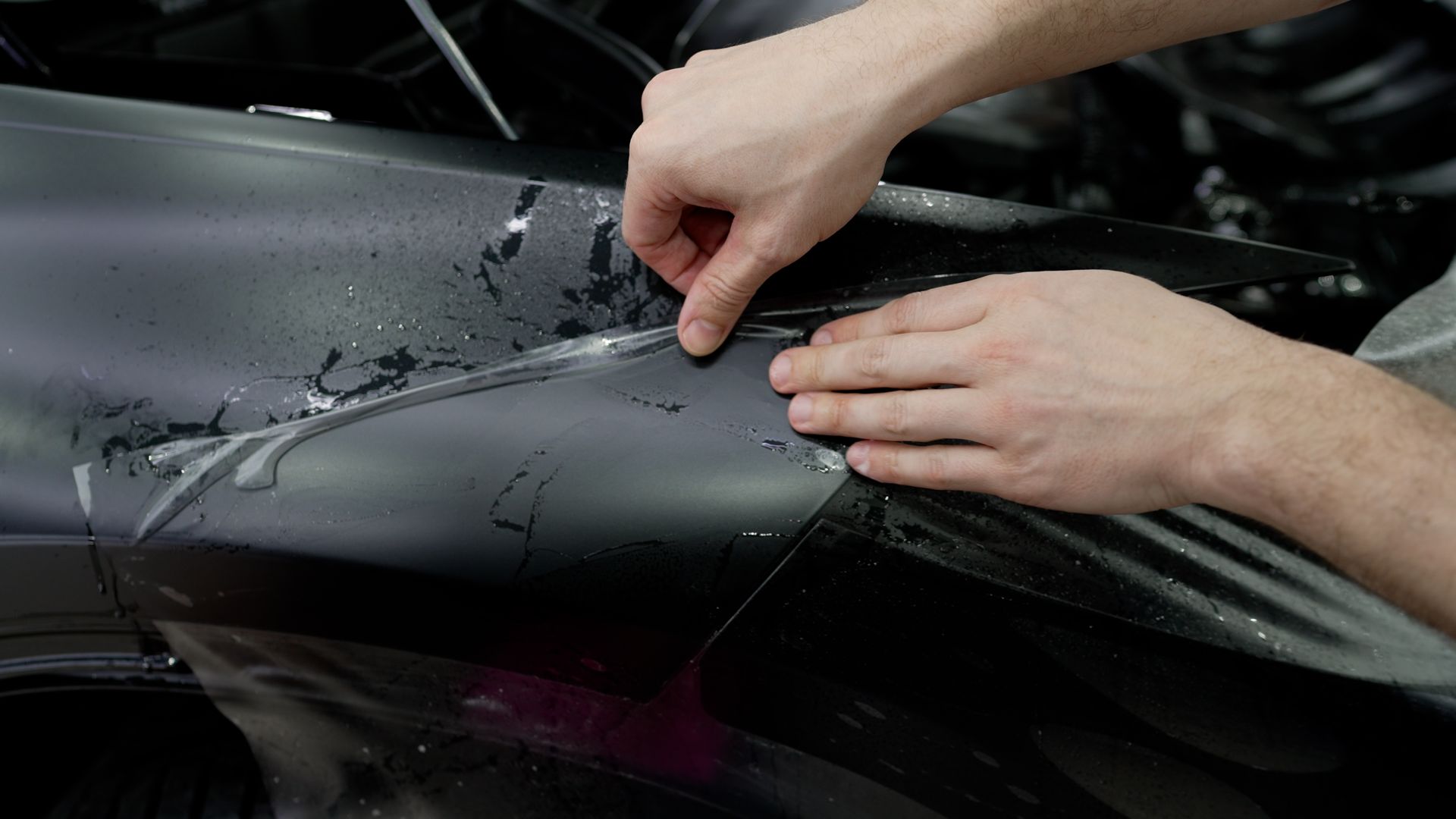 Hands peeling protective film from a dark car surface, likely wrapping or detailing.