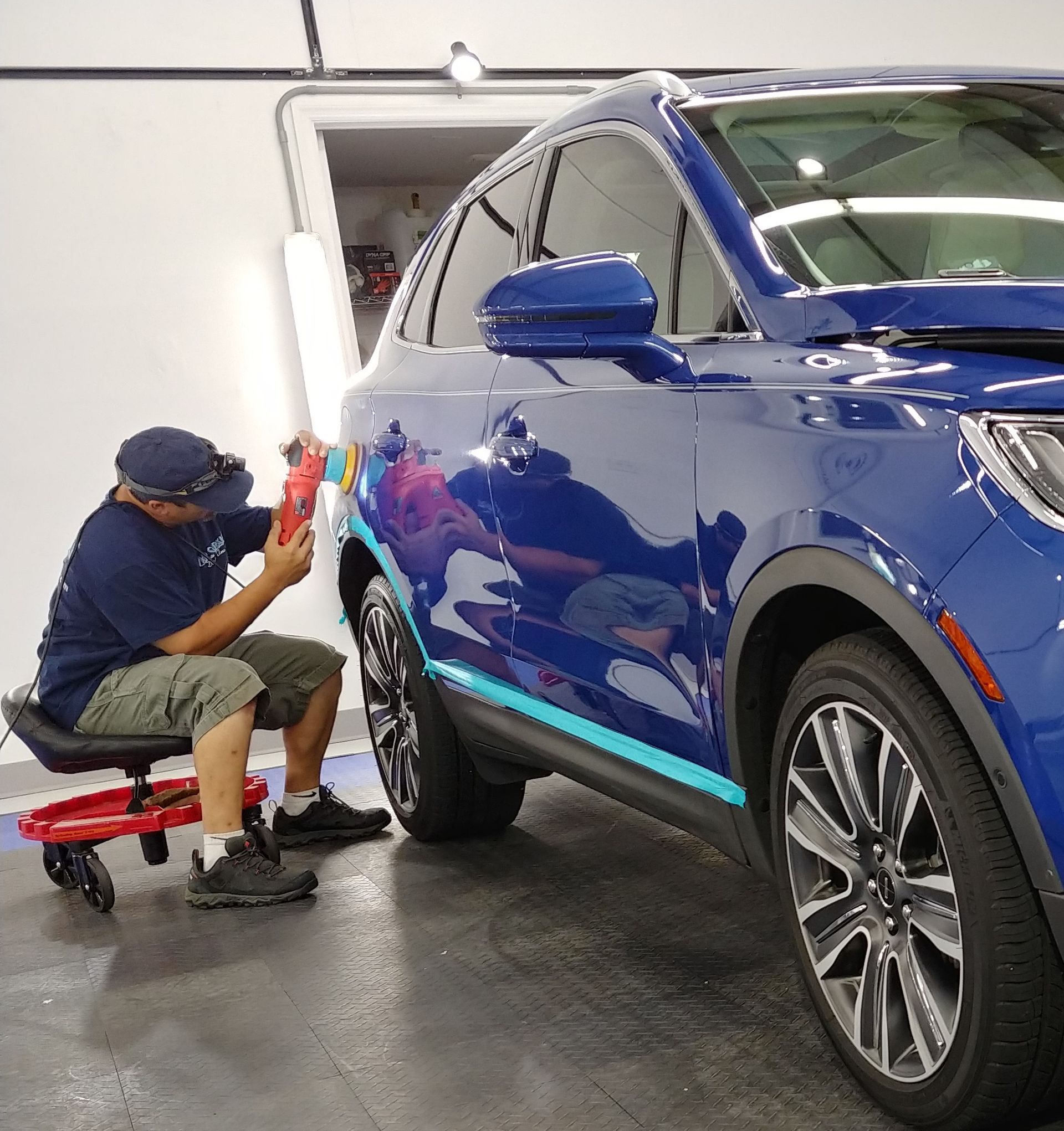 Man working on a blue car in a garage, using a light to inspect for imperfections.