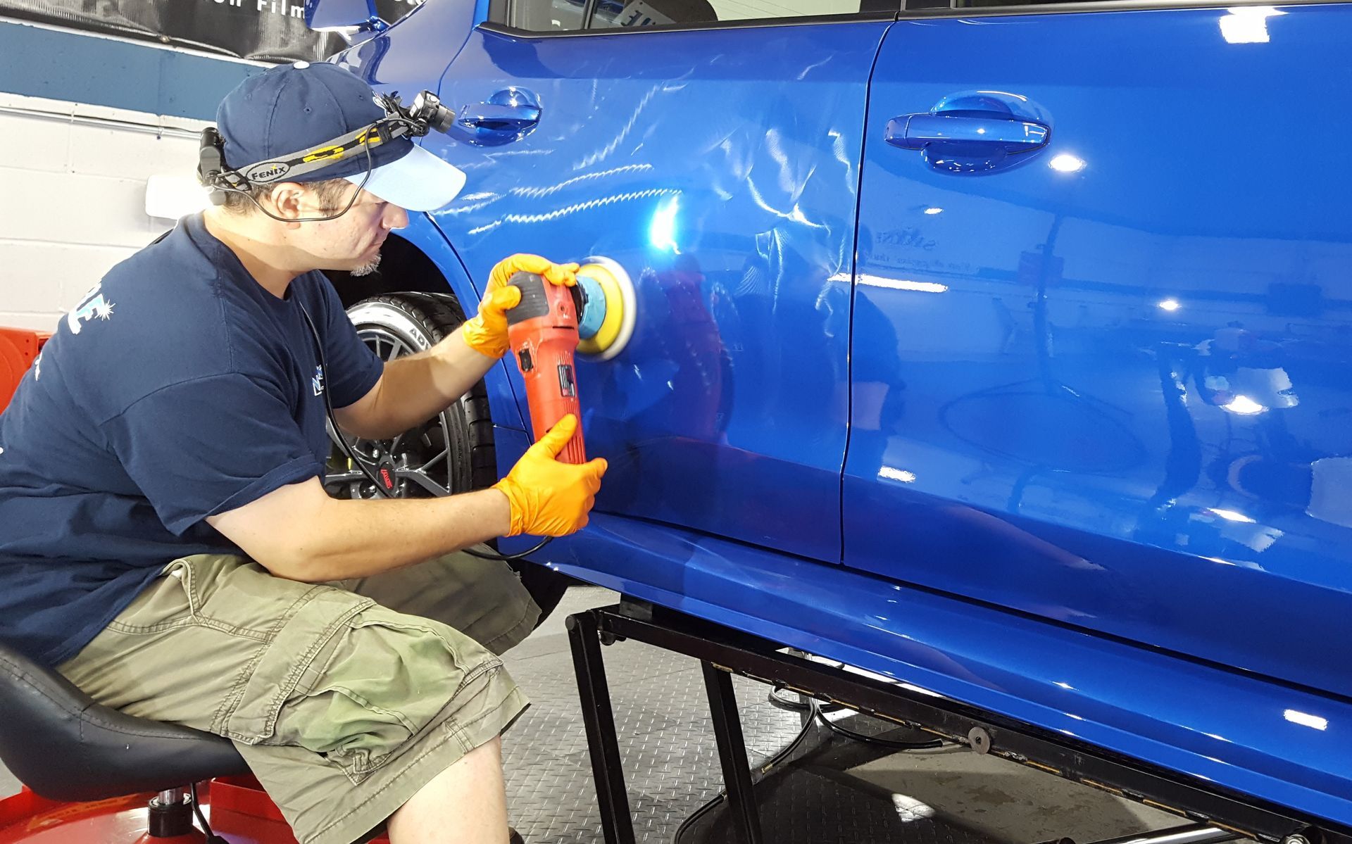 Man polishing blue car door with an orange buffer, wearing gloves and headlamp.