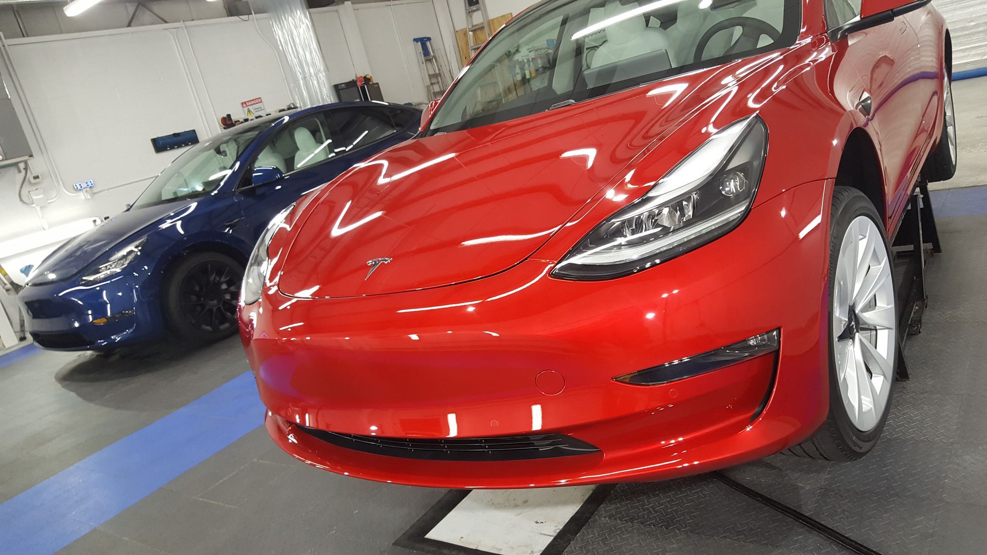 Red Tesla car in the foreground, blue Tesla car in the background, both parked in a garage.