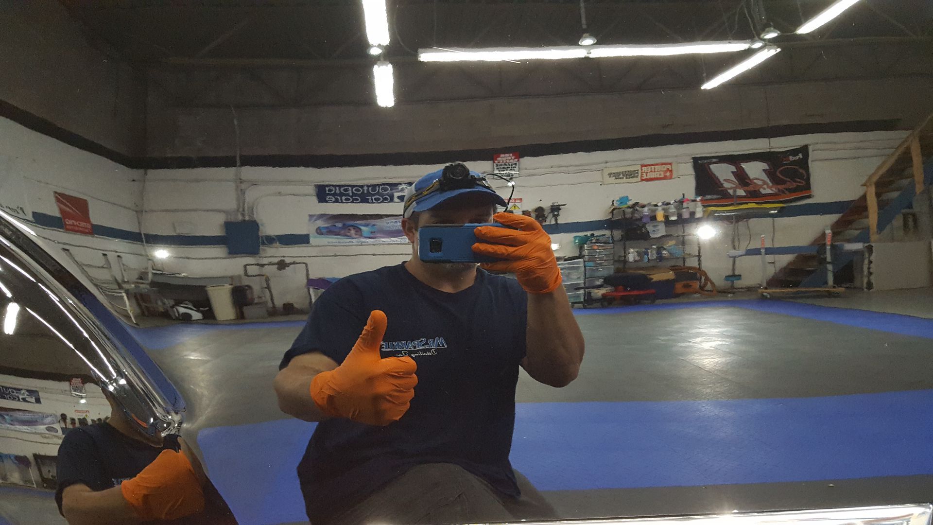 Man wearing gloves in auto shop, taking selfie, giving thumbs up.