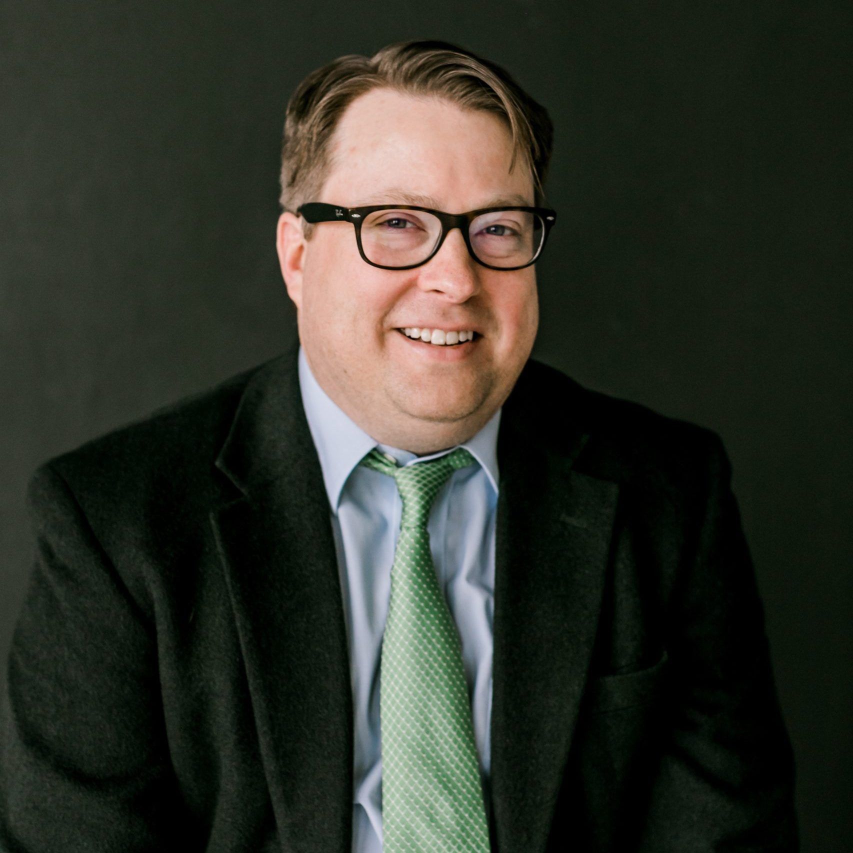 Cullen Mcmahon with glasses wearing a suit and green tie, smiling at the camera. Dark background.