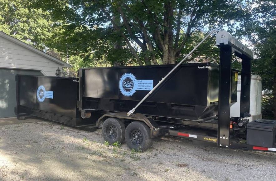 A dumpster on a trailer is parked in front of a garage.