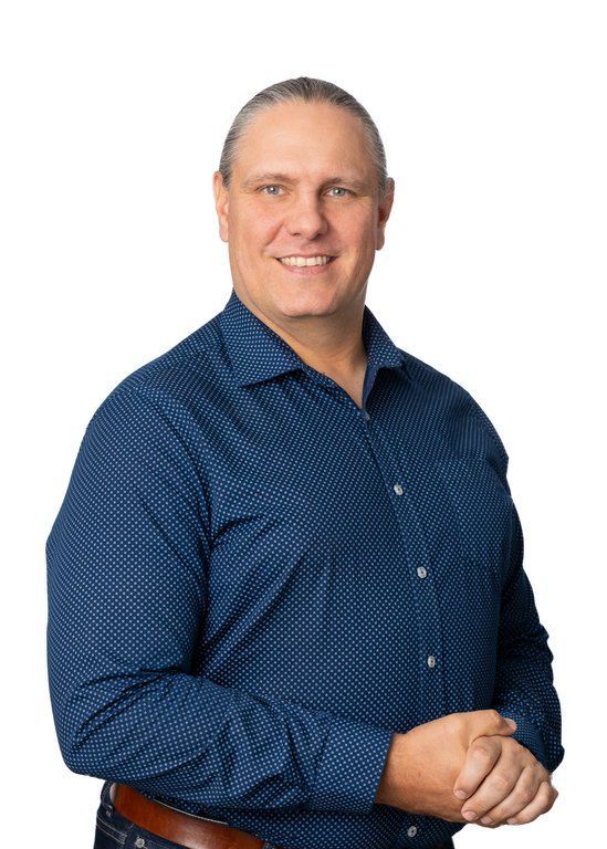 A man in a blue shirt is standing with his hands folded and smiling.