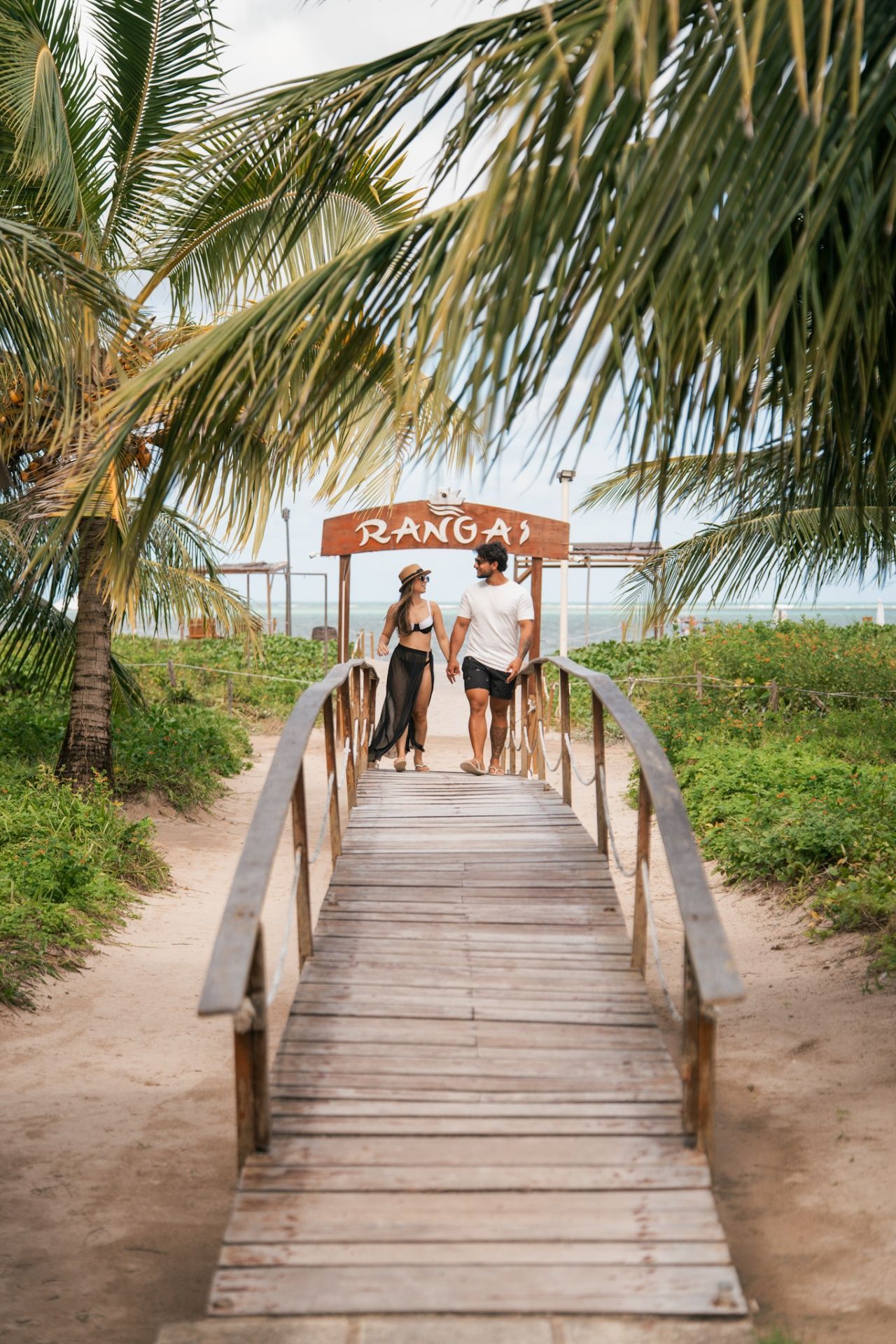 Um casal caminha de mãos dadas por uma ponte de madeira que leva à entrada de uma praia tropical com a placa "RANGAI", sob palmeiras.