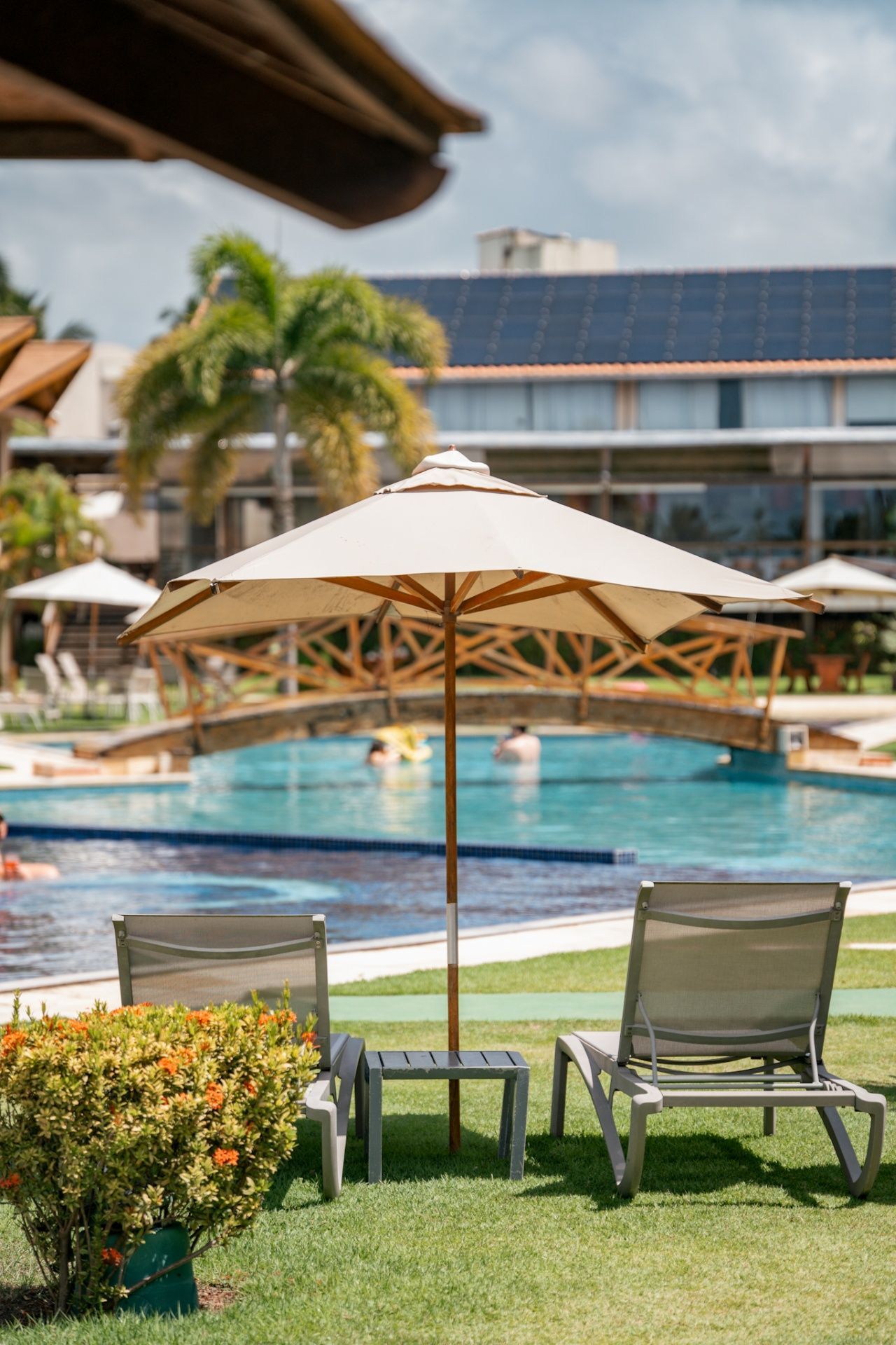 Duas espreguiçadeiras com um guarda-sol à beira da piscina num dia ensolarado, com uma pequena passarela sobre a água e o hotel ao fundo.