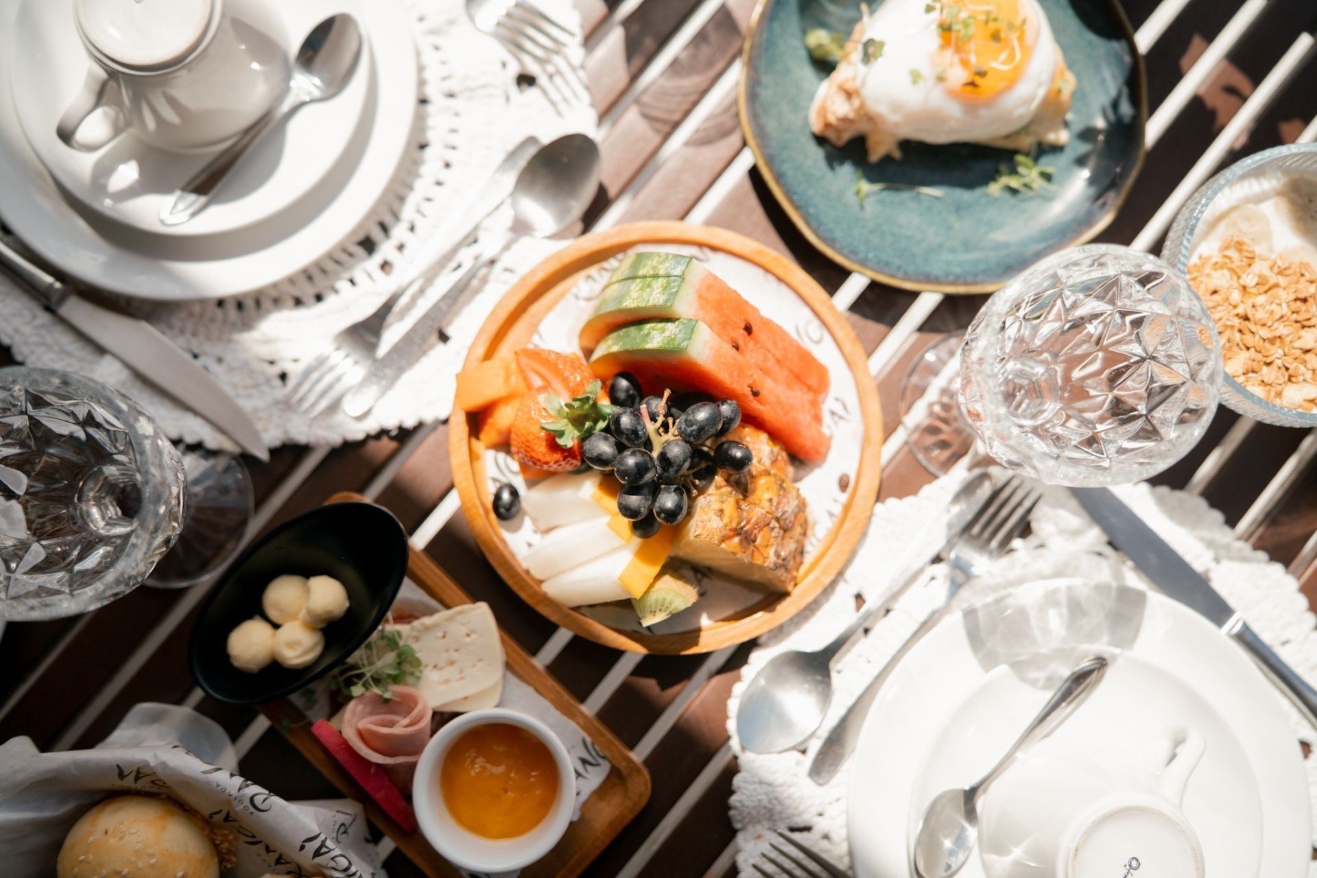 Vista de cima de uma mesa de café da manhã posta com frutas, pão, manteiga, ovos e bebidas, dispostos sobre uma mesa listrada branca.