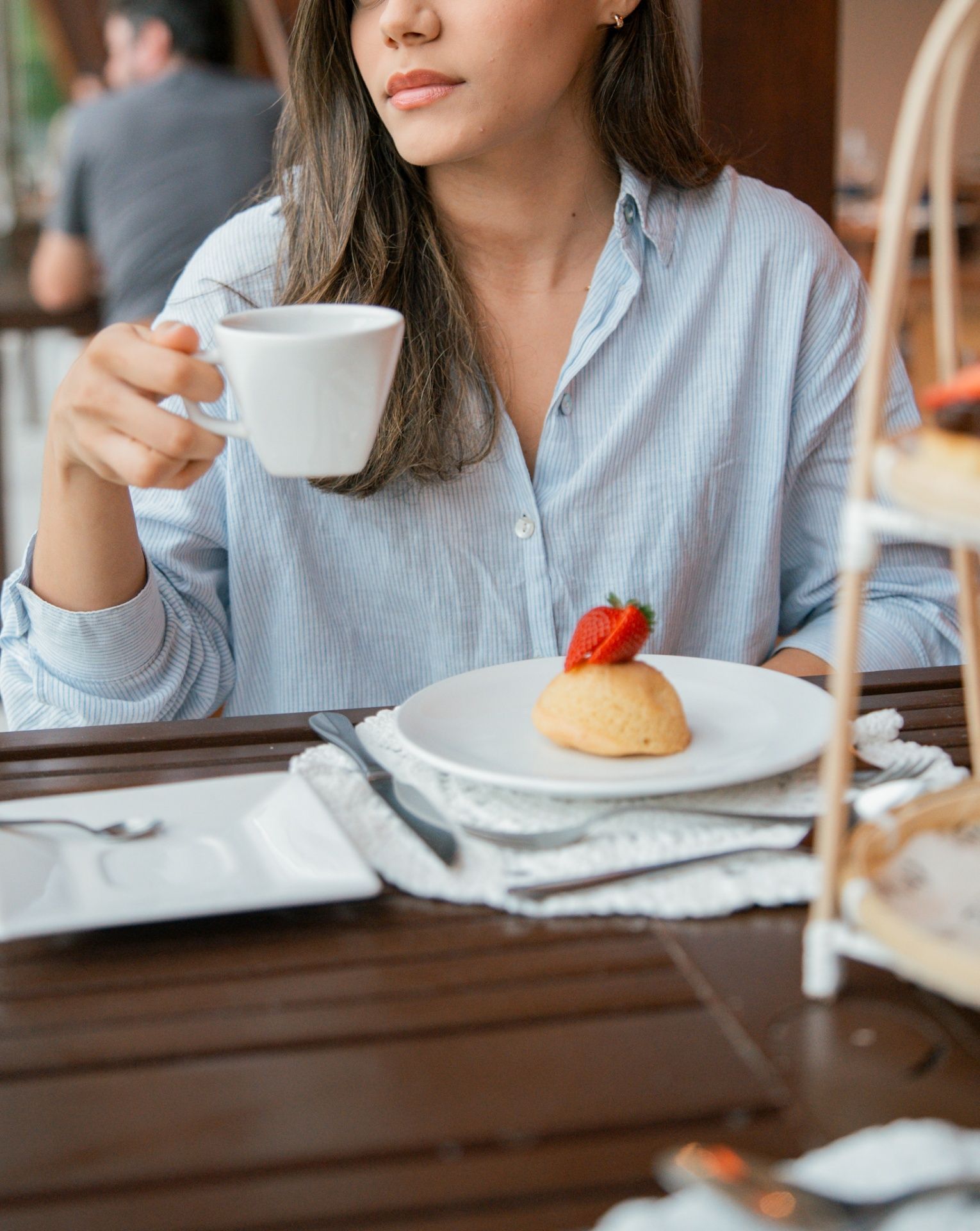 Uma pessoa vestindo uma camisa azul-clara segura uma xícara de café branca sobre uma mesa de madeira com um pequeno bolo decorado com um morango.