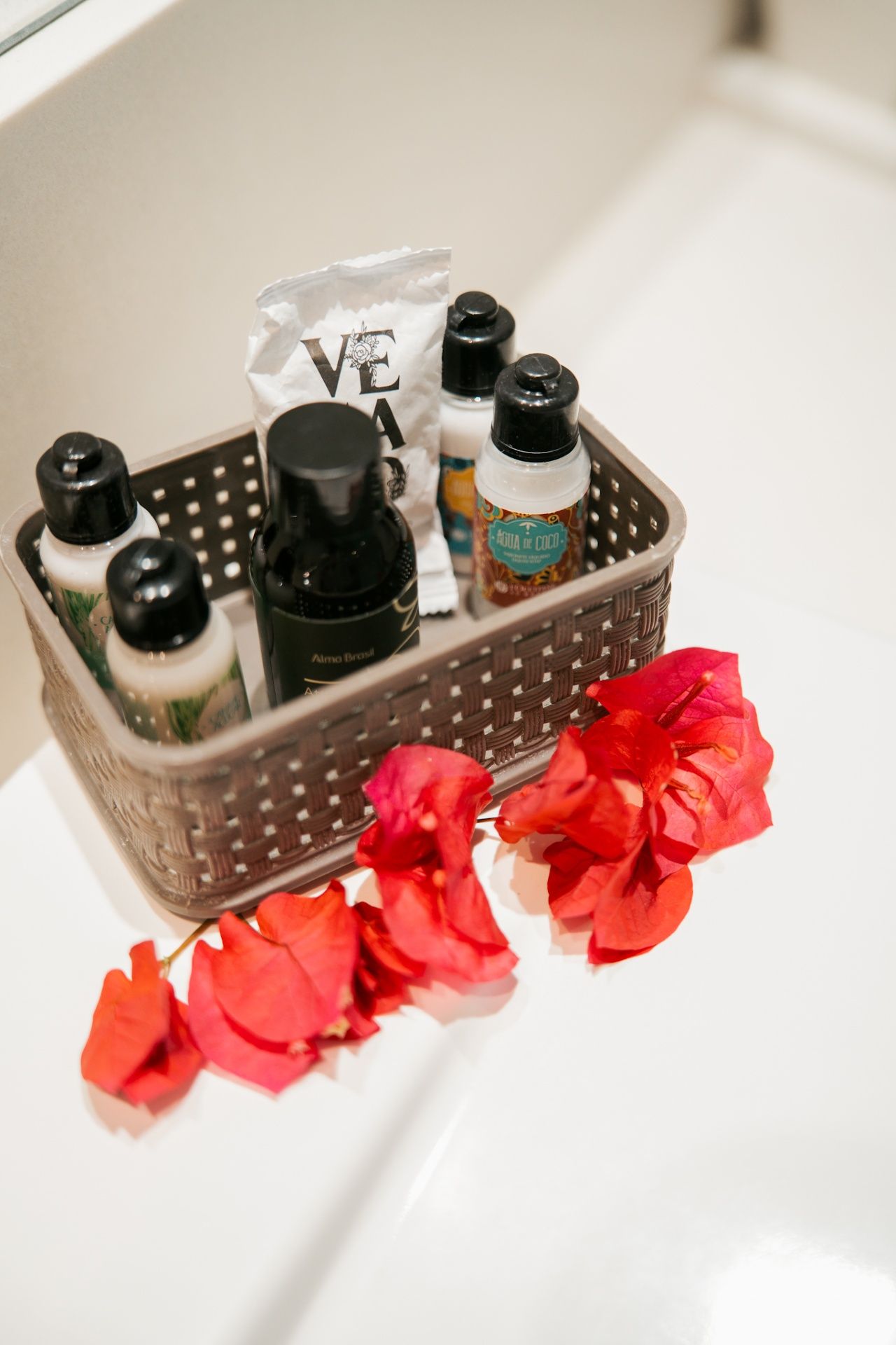 Uma cesta de plástico marrom contendo pequenos artigos de higiene pessoal de hotel repousa sobre uma superfície branca, adornada com flores rosa vibrantes.