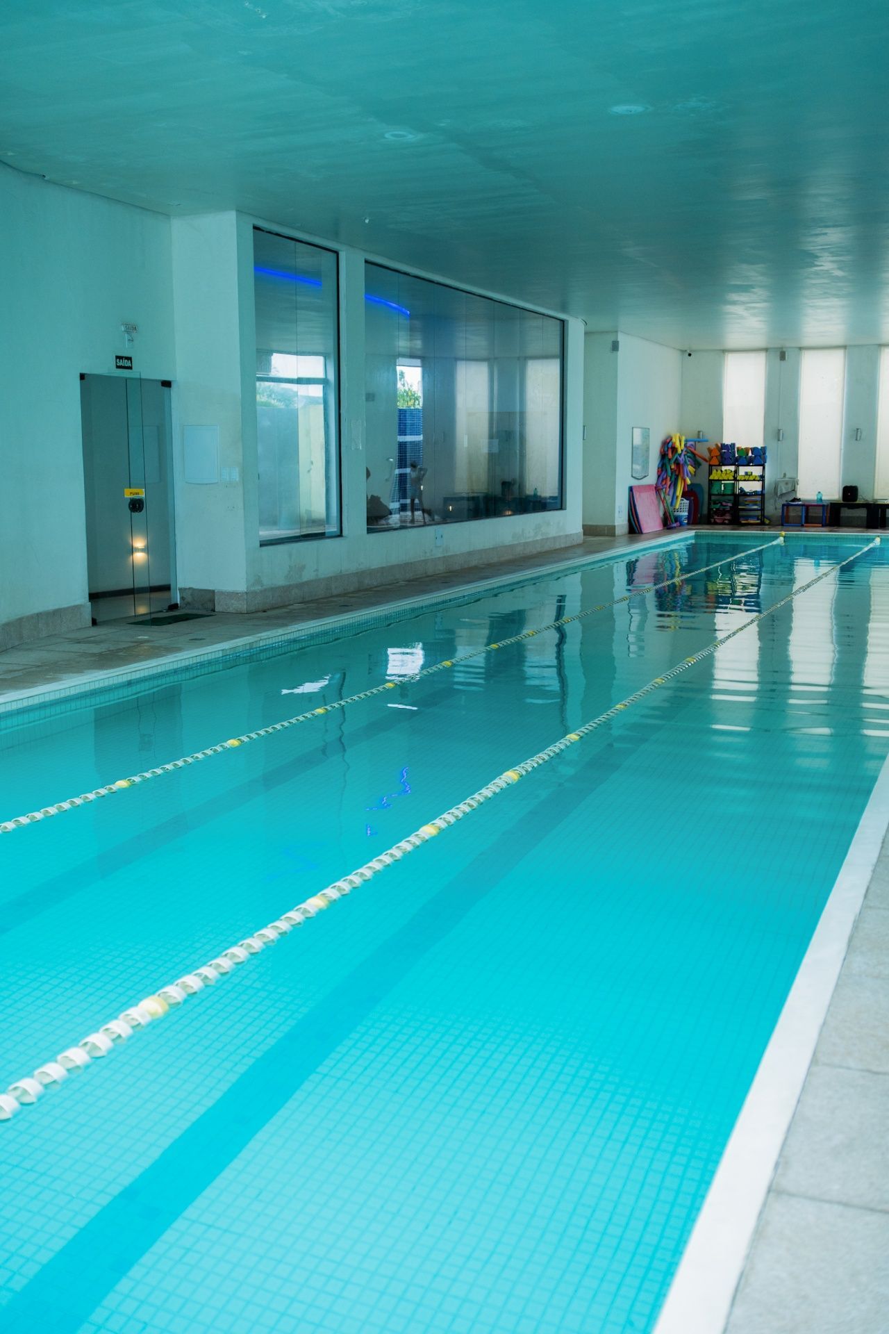 Uma piscina coberta com água azul e linhas demarcadoras de raias, vista do deck lateral.