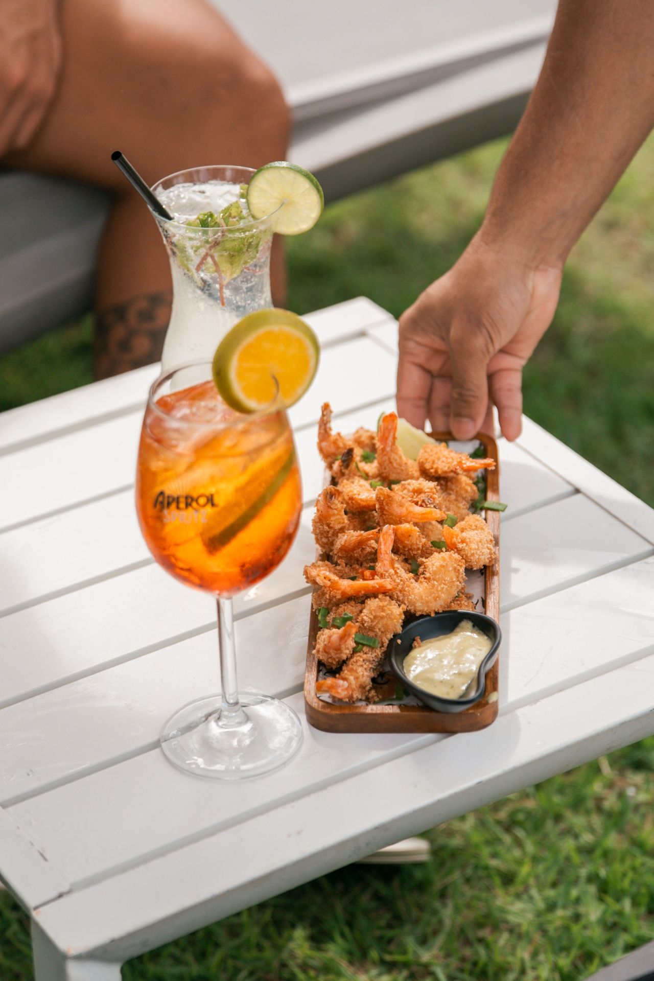 Uma mão coloca uma travessa de madeira com camarões fritos ao lado de um Aperol Spritz e um coquetel transparente em uma mesa branca ao ar livre.