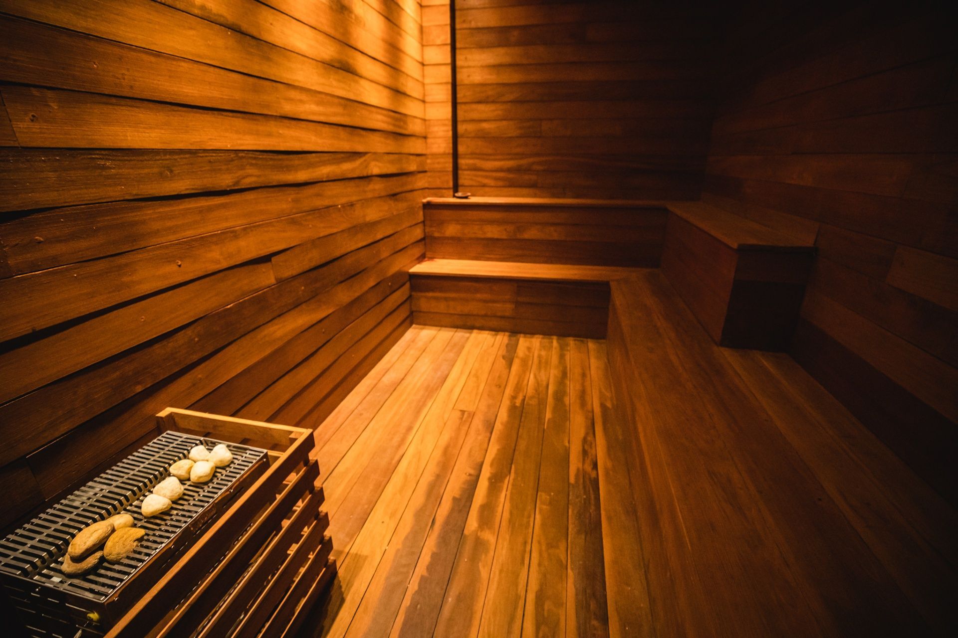 Uma sauna aconchegante e com iluminação suave, revestida com painéis de madeira horizontais, bancos de madeira embutidos e um aquecedor preenchido com pedras.
