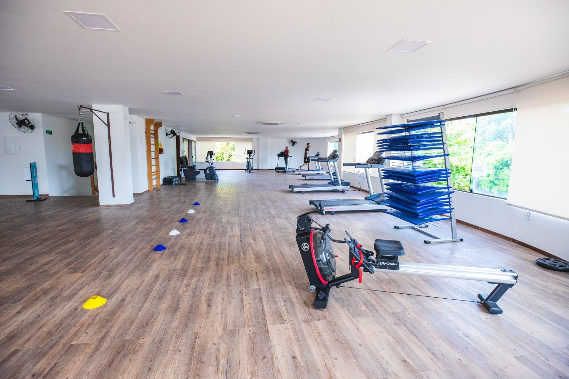 Interior de ginásio com piso de madeira, máquina de remo, saco de pancadas, esteiras e um suporte de pesos azul.