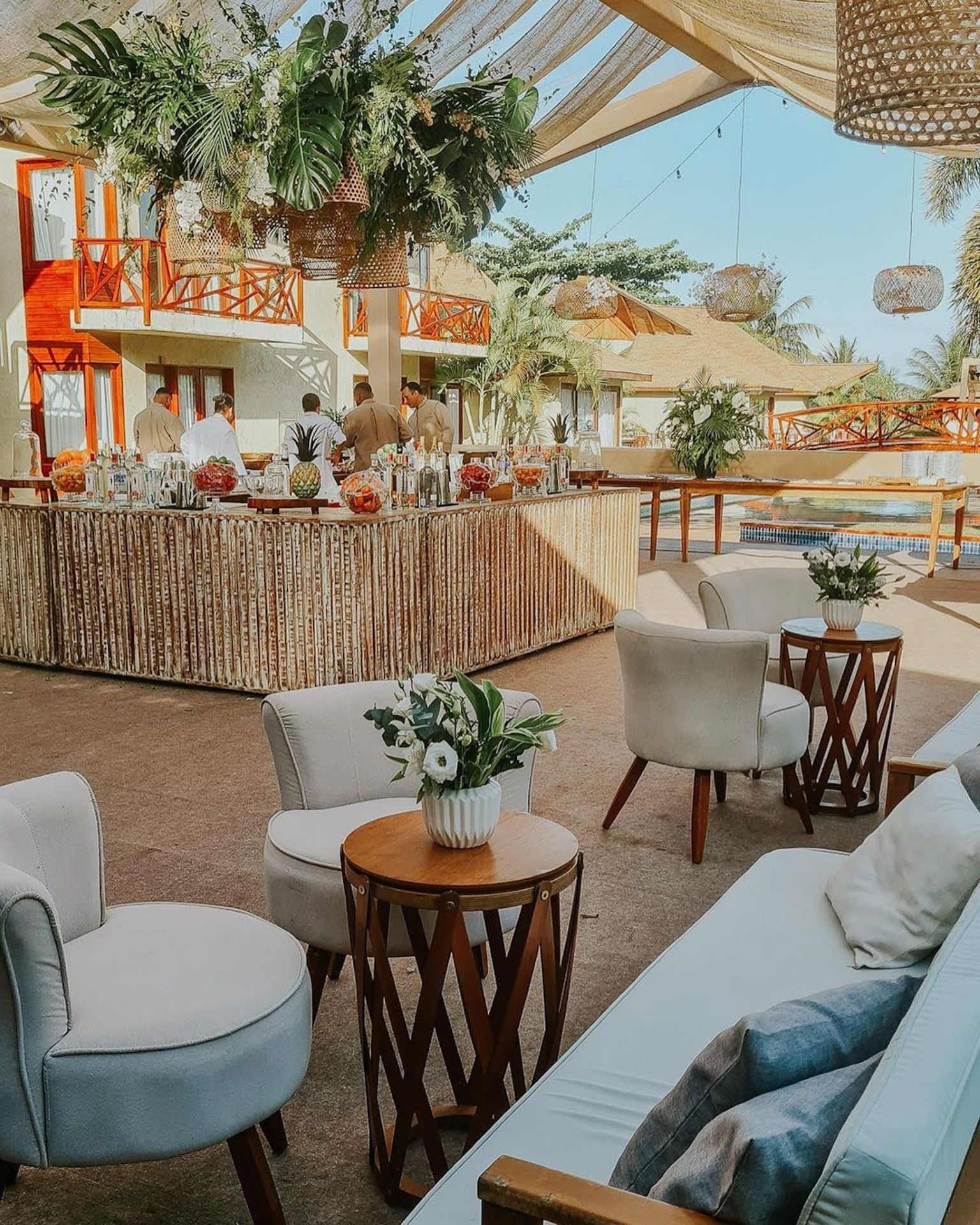 Uma área de estar externa com cadeiras cor creme, pequenas mesas de madeira e um bar com temática tropical, situada em um resort.