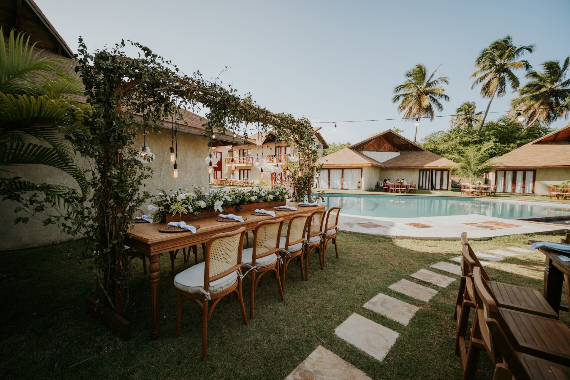 Uma mesa de jantar de madeira posta para um evento em um gramado ao lado de uma piscina, à sombra de um caramanchão coberto de vegetação.