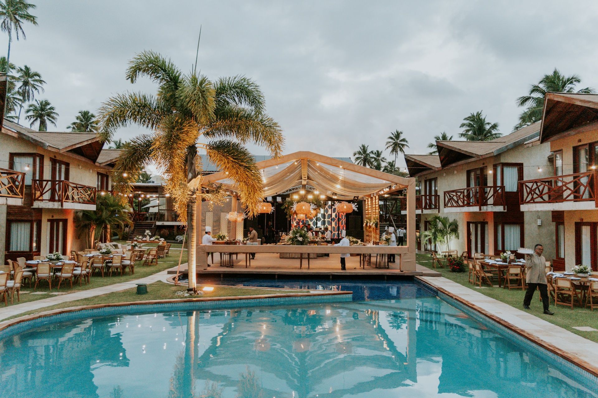 Um espaço ao ar livre com um palco decorado sob uma tenda, ladeado por edifícios de dois andares e uma piscina em primeiro plano.
