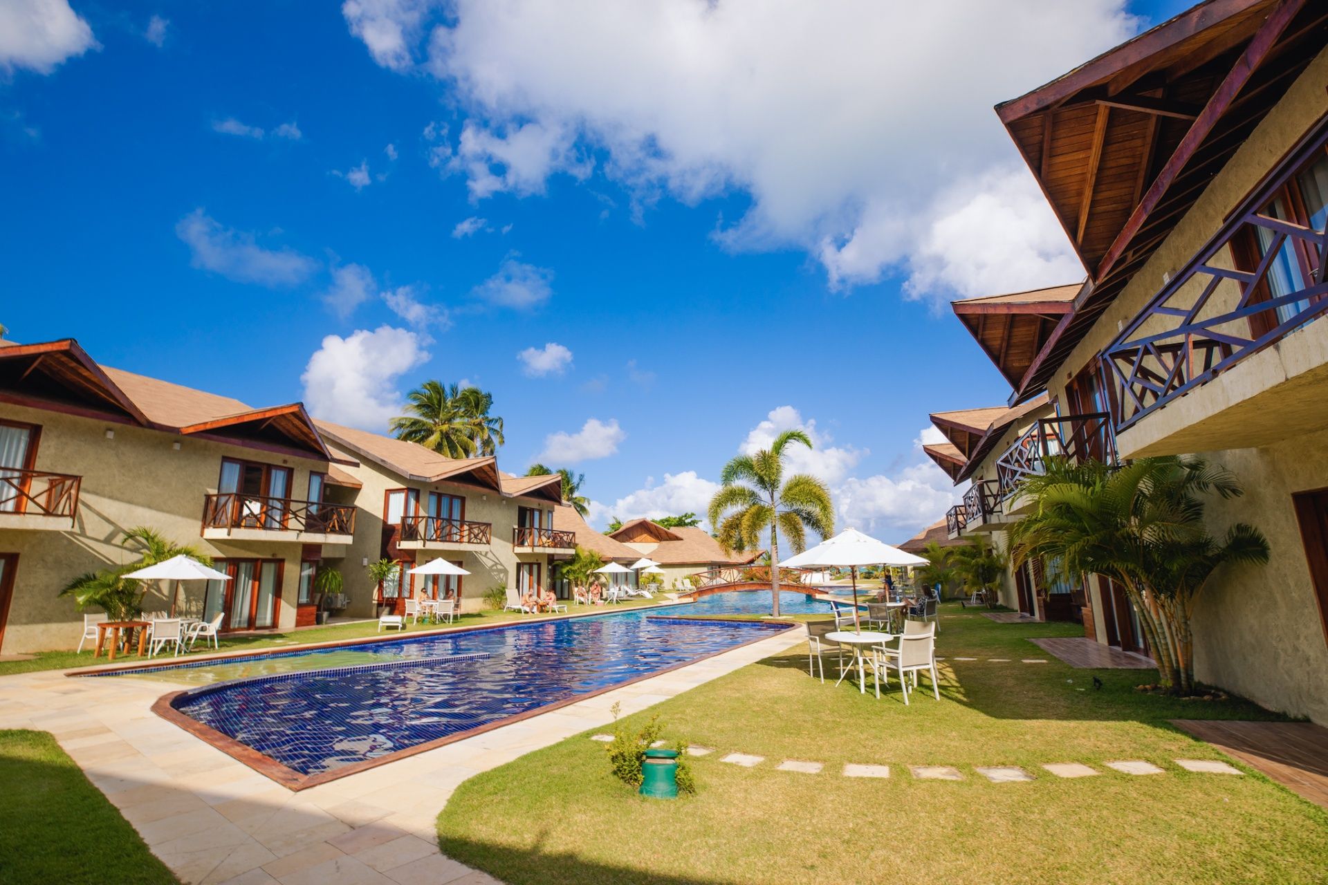 Uma cena ensolarada de um resort com uma piscina retangular cercada por prédios baixos, palmeiras e mesas ao ar livre.