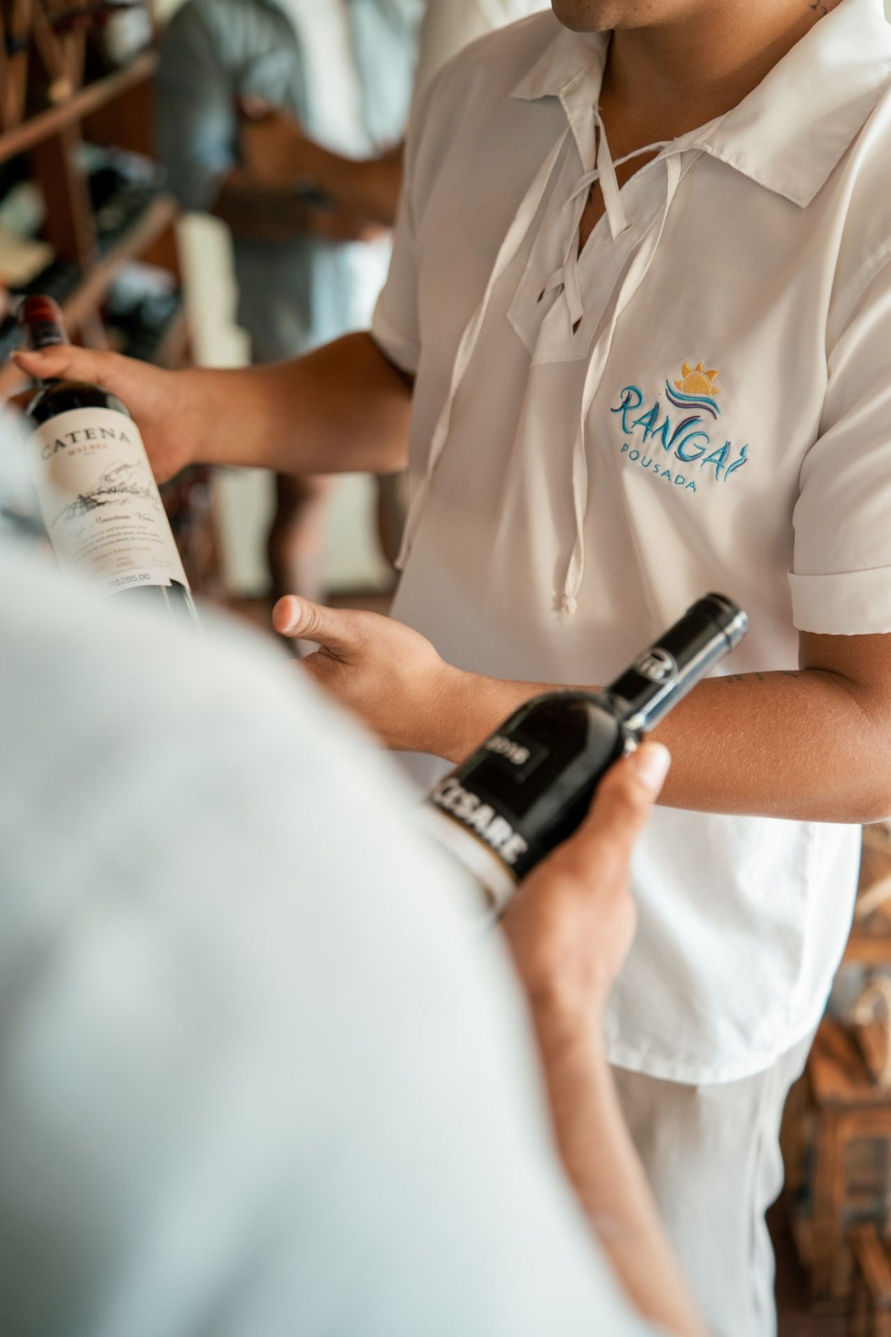 Uma pessoa vestindo uma camisa branca de colarinho, segurando uma garrafa de vinho, gesticula para outra pessoa que segura uma garrafa de vinho preta.