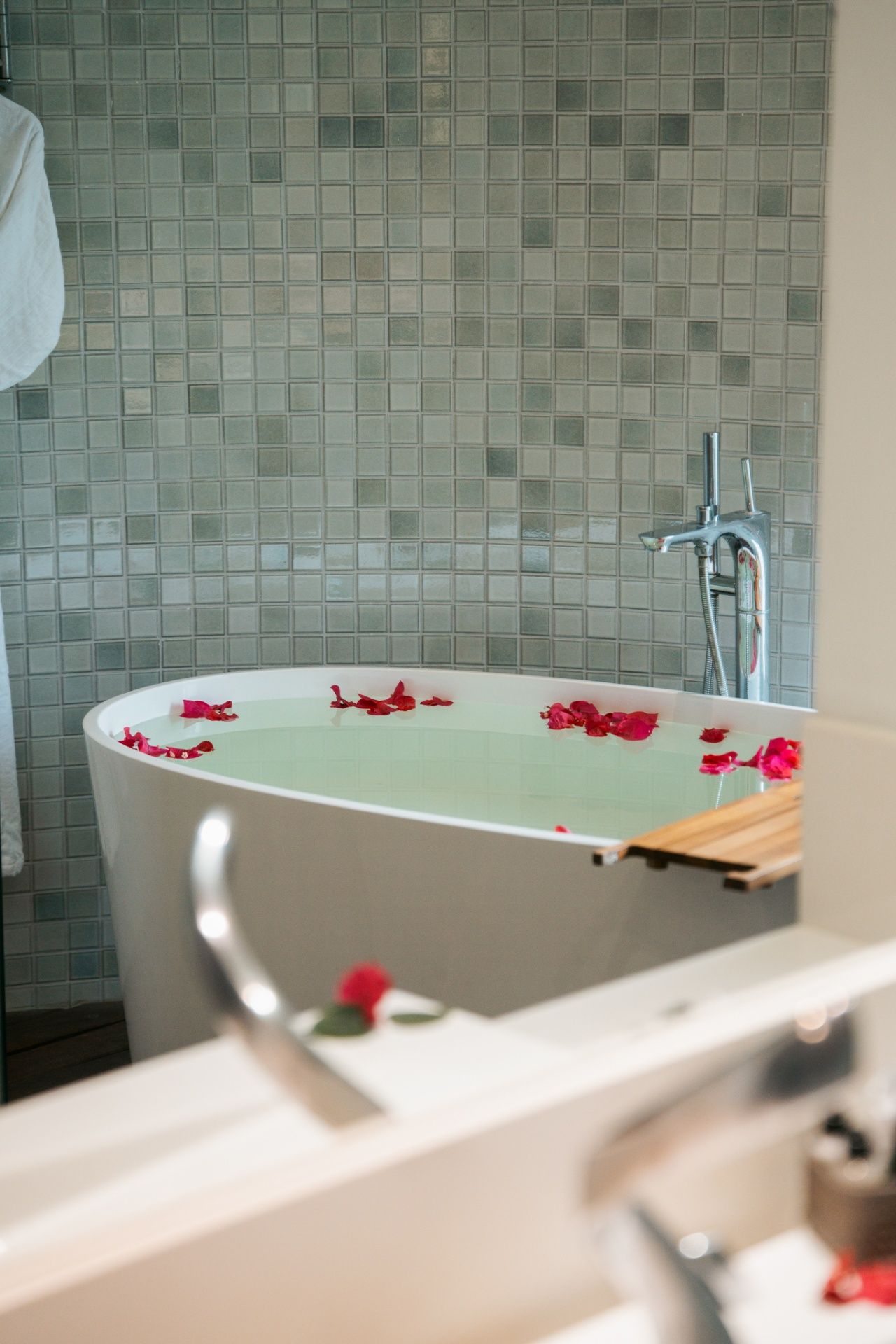Uma banheira branca, oval e independente, cheia de água e com pétalas de rosas vermelhas espalhadas contra uma parede revestida com azulejos cinza texturizados.