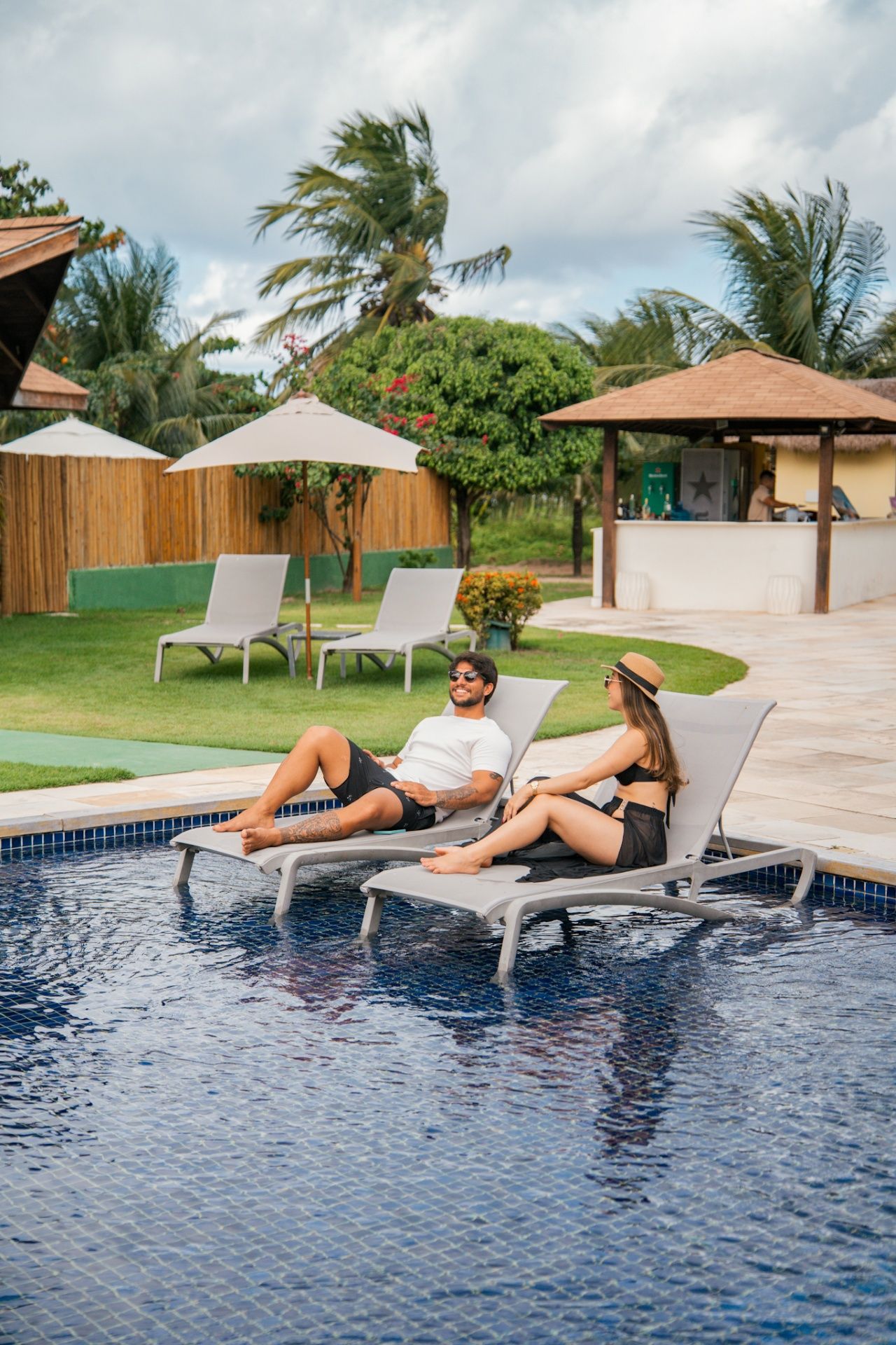 Um casal relaxa em espreguiçadeiras parcialmente submersas em uma piscina em um resort com bar e palmeiras nas proximidades.