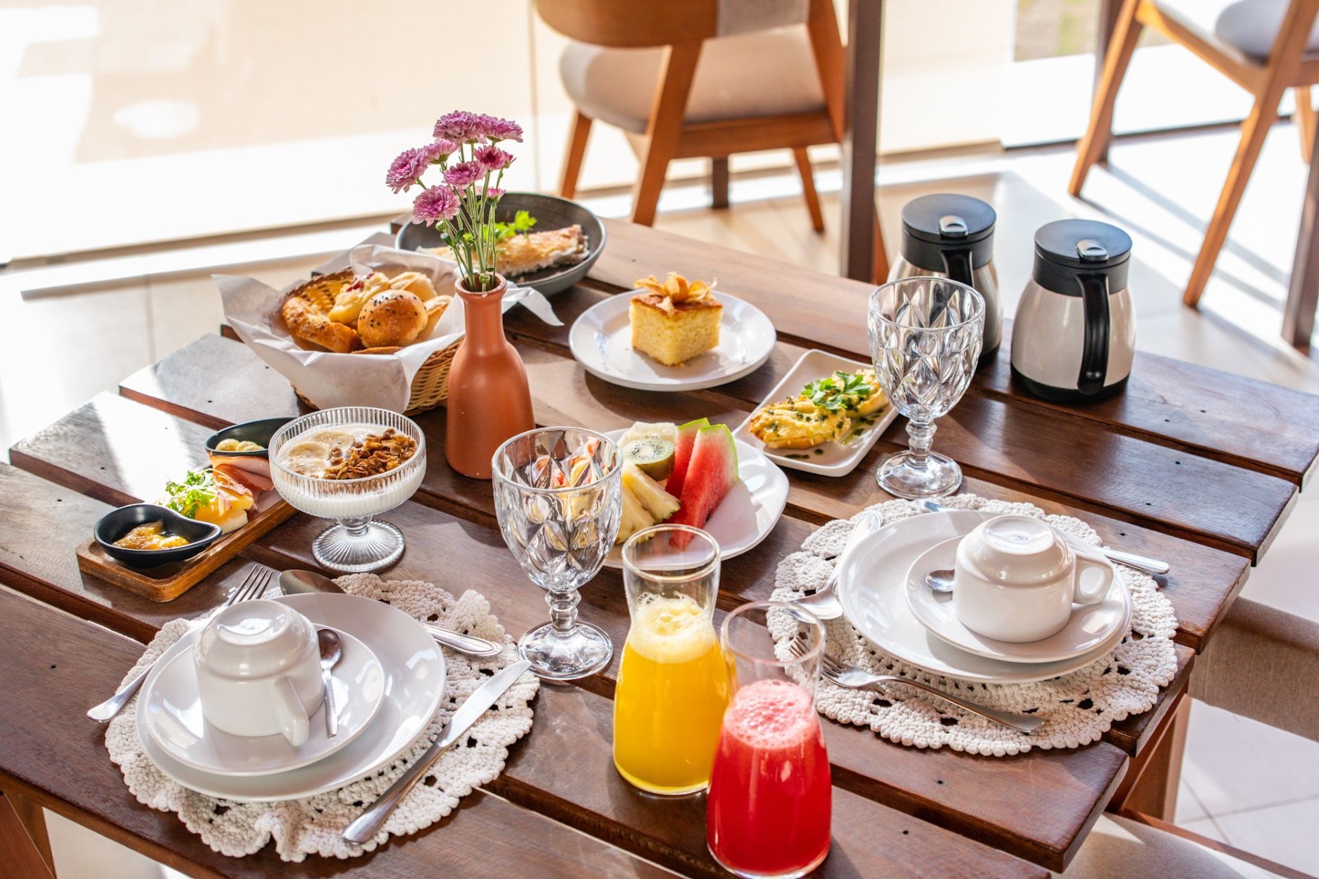 Uma mesa de café da manhã de madeira posta para dois com doces, frutas frescas, iogurte, suco e café em uma sala bem iluminada.