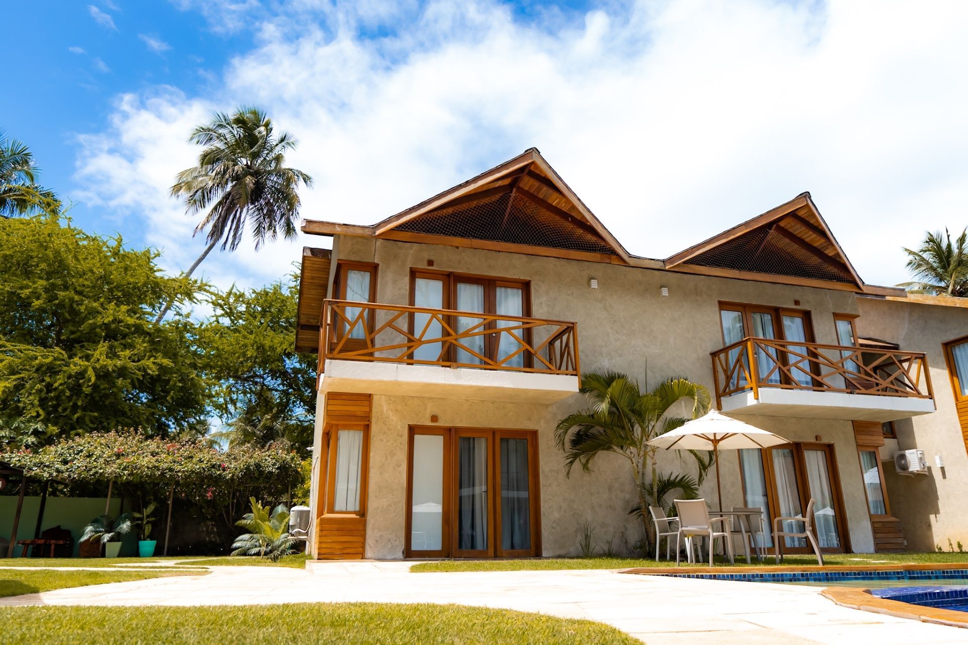 Uma casa de dois andares com paredes de estuque bege, varandas de madeira e telhados pontiagudos, situada junto a uma piscina sob um céu azul.