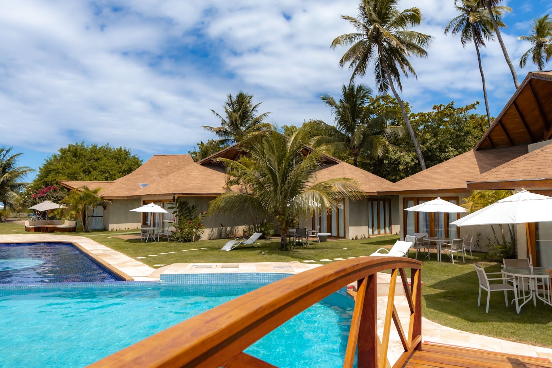 Resort tropical com piscina, ponte de madeira, guarda-sóis brancos e edifícios rodeados por palmeiras exuberantes.