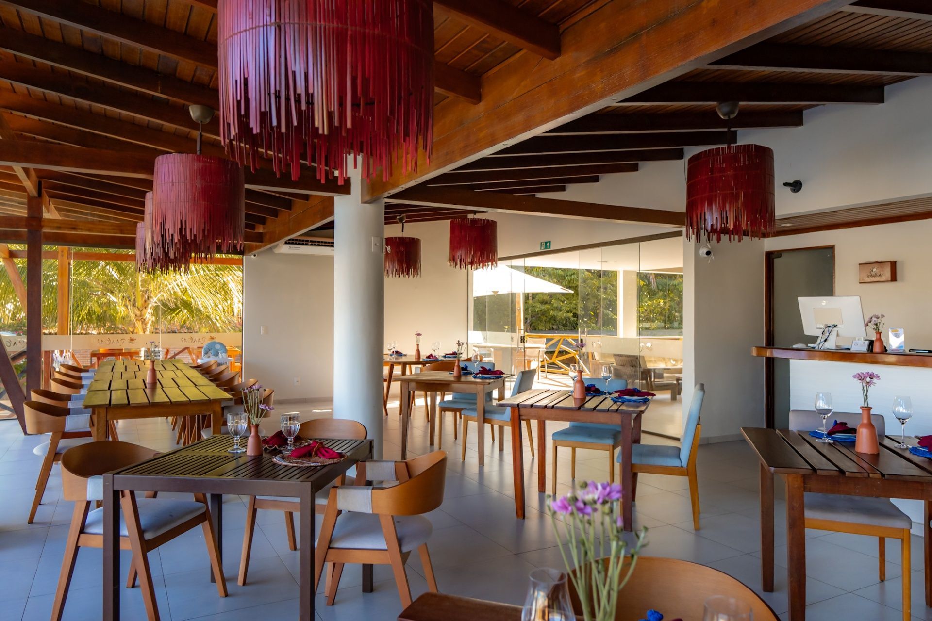 Uma área de jantar ao ar livre com mesas e cadeiras de madeira, e grandes luminárias pendentes vermelhas com franjas penduradas no teto.