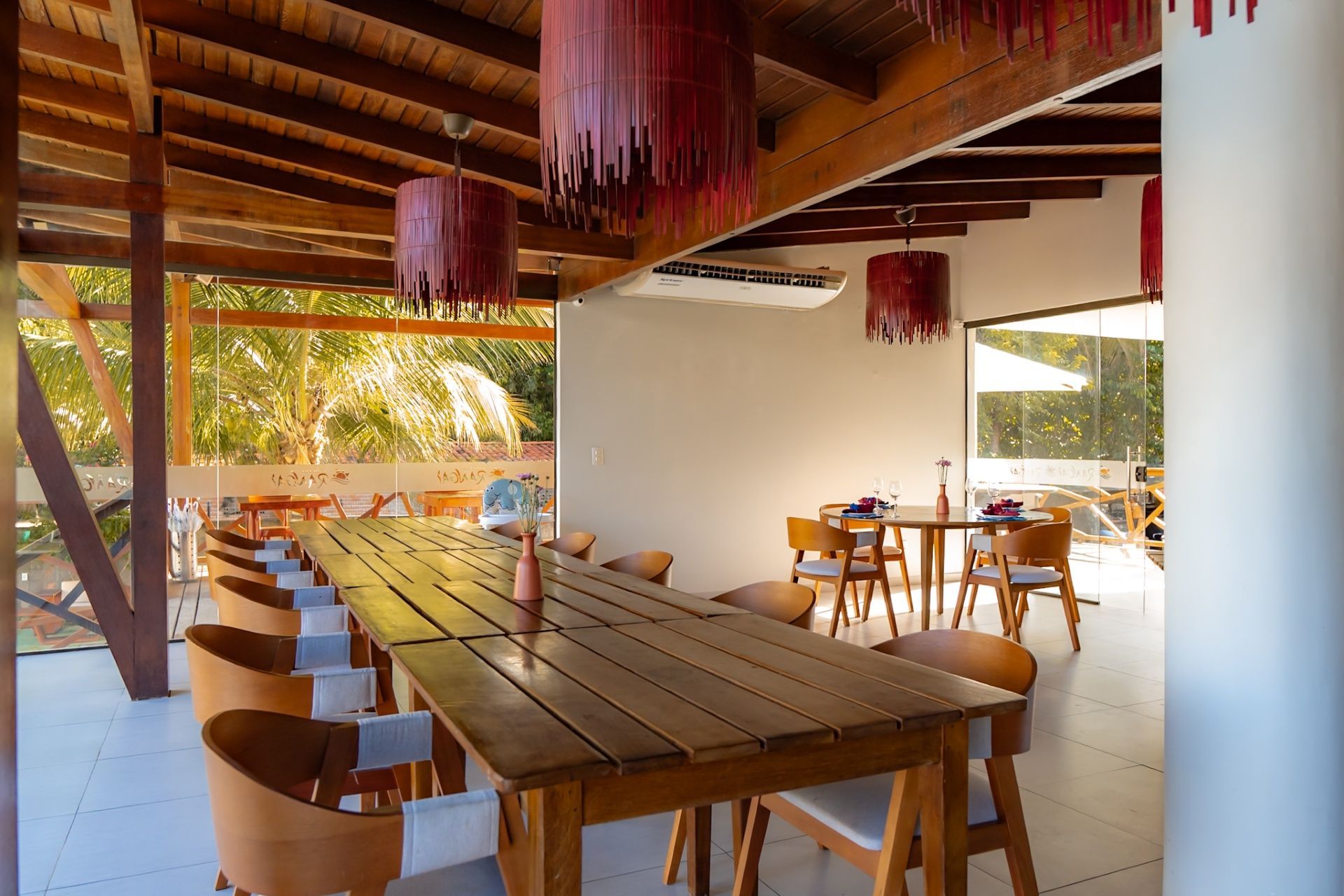 Mesa comprida de madeira em uma área de jantar ao ar livre com luminárias rosa penduradas e vistas panorâmicas do exterior.