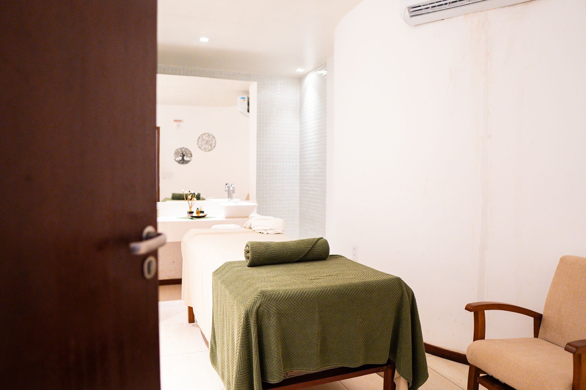Uma sala de massagem com duas macas cobertas com mantas verdes, visível através de uma porta de madeira aberta.