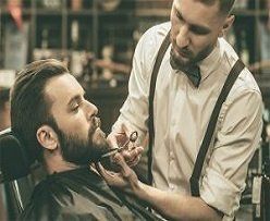 Barber trimming a customer's beard in a barbershop. The barber wears suspenders and a bow tie.
