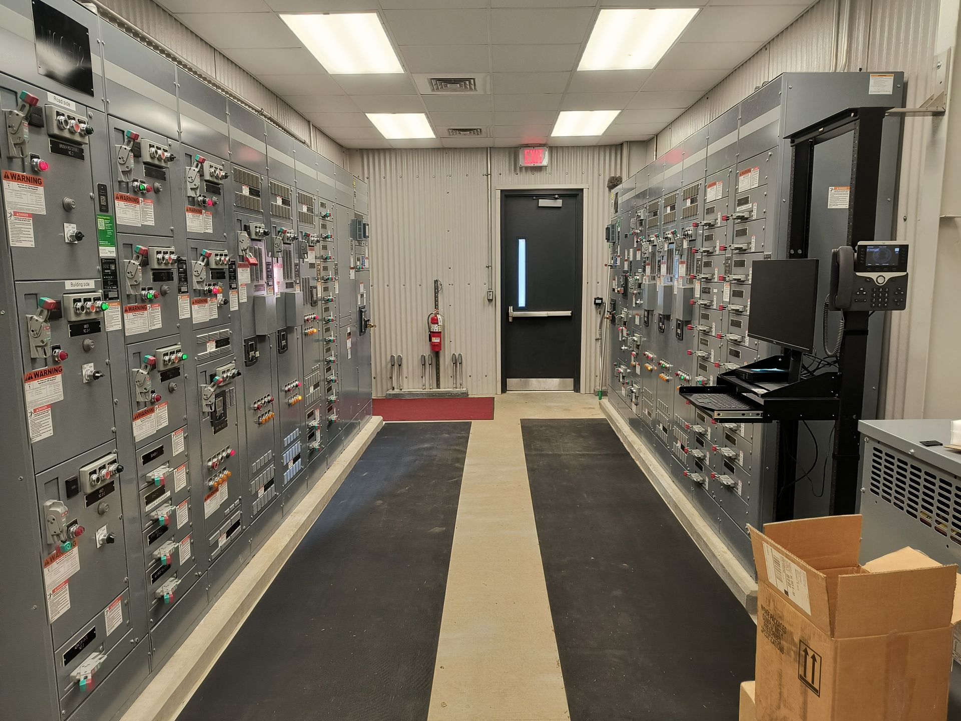 Electrical control room with rows of gray panels, overhead lights, and a doorway at the far end.