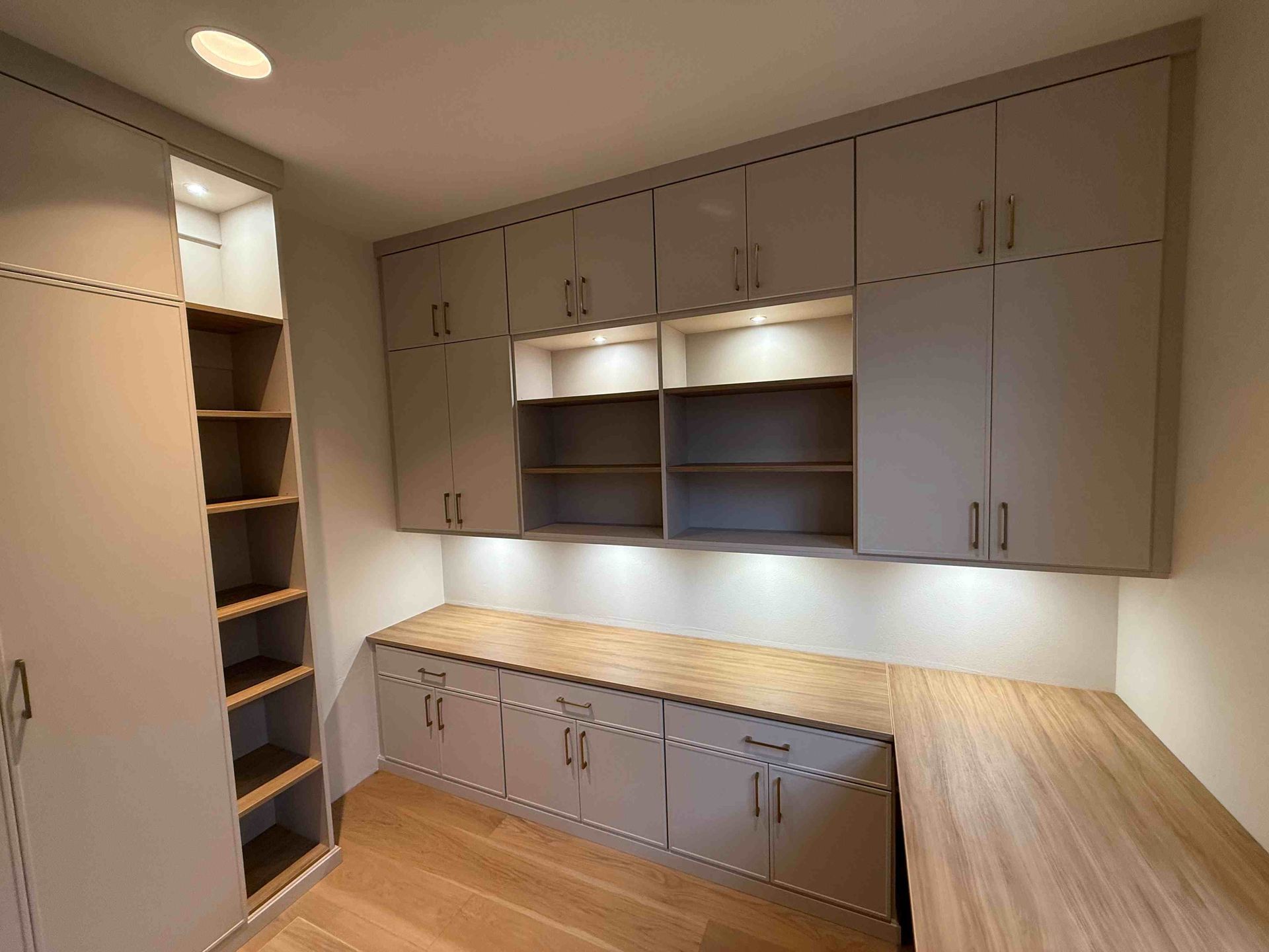 A modern office with gray cabinets, wooden desk, and shelves. Soft lighting and window.