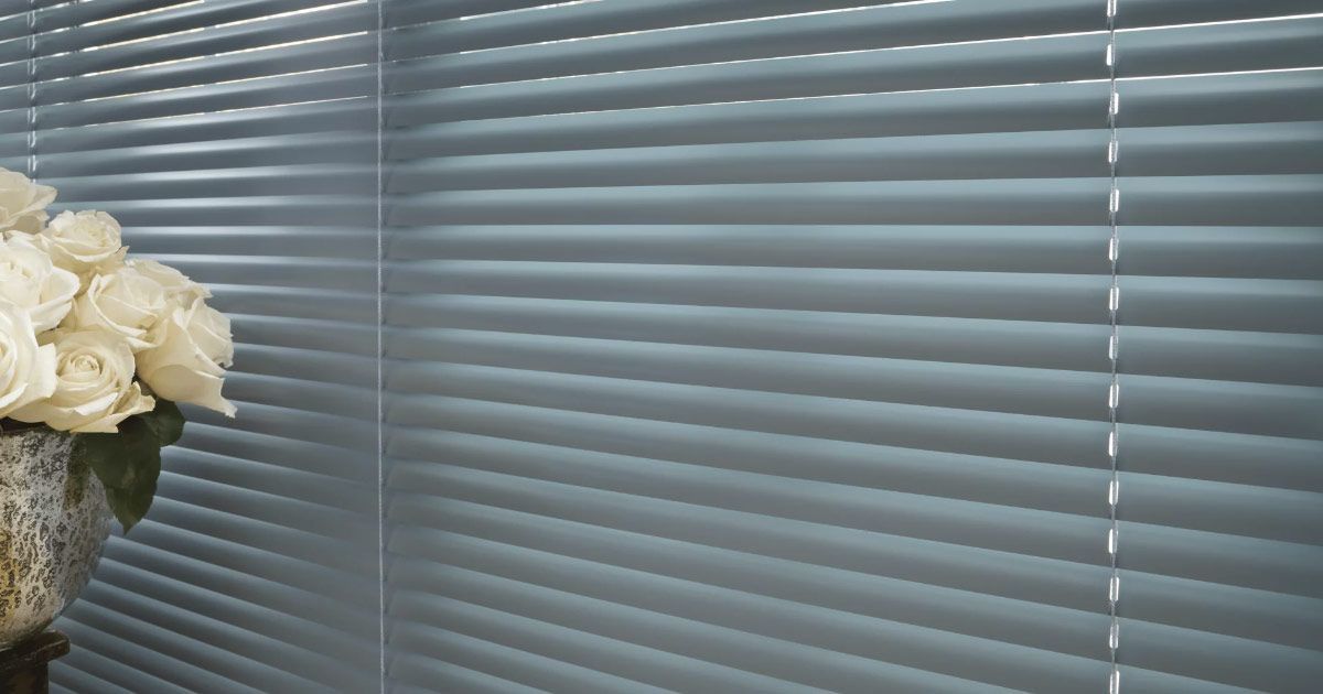 A vase of white roses is sitting in front of a window with blinds.