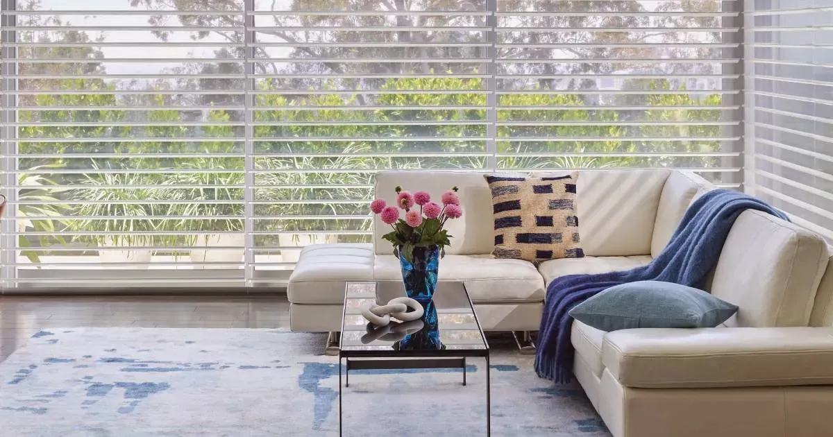 A living room with sheer shades, a couch , coffee table , and a vase of flowers.