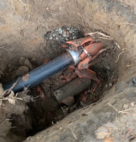 Broken sewer pipes in an excavated hole, with a black plastic pipe and broken terracotta pipe