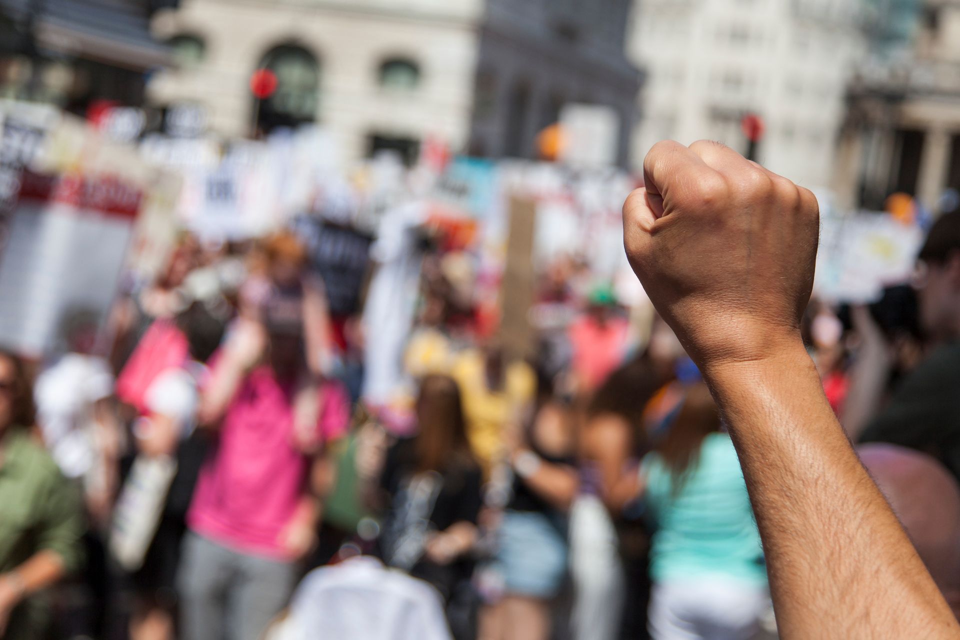 Person's fist raised in a crowd of people at a protest, blurred background of signs and buildings.