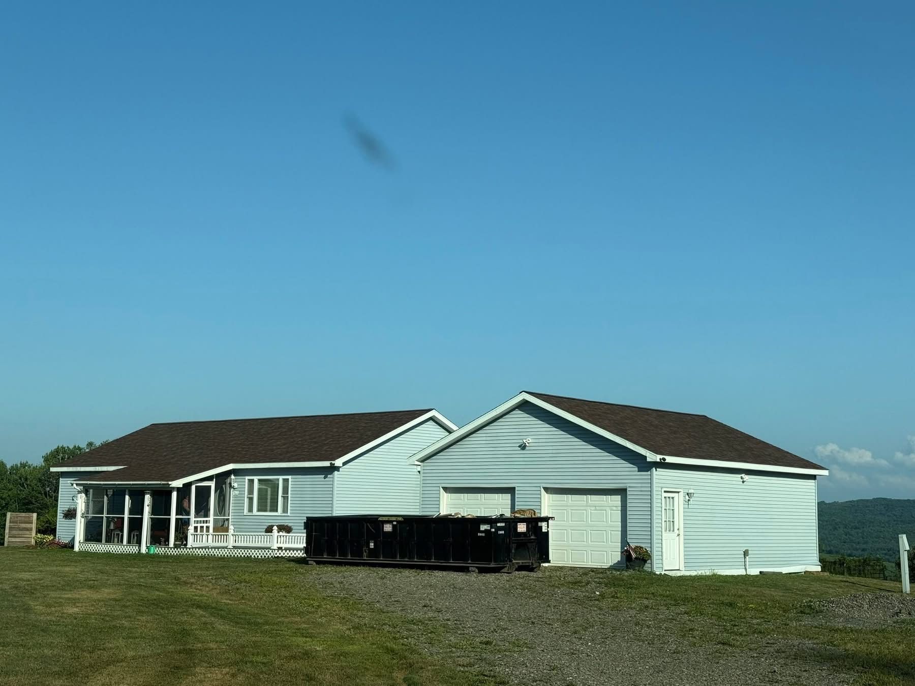 Blue house and garage with brown roofs, a dumpster in front, on a grassy hill under a blue sky.