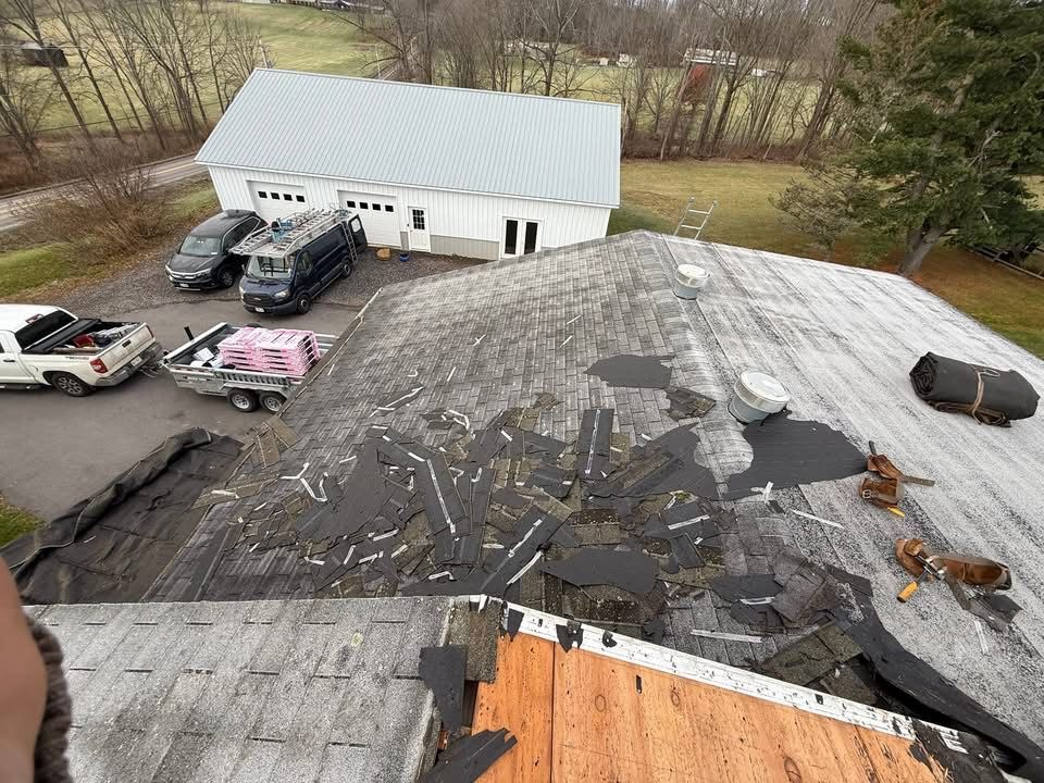 Roof with exposed wood, damaged shingles, and construction vehicles in a rural setting.