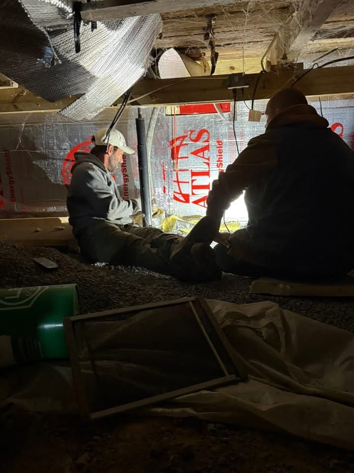Two people installing insulation under a building. Low light, gravel floor, Atlas logo visible.