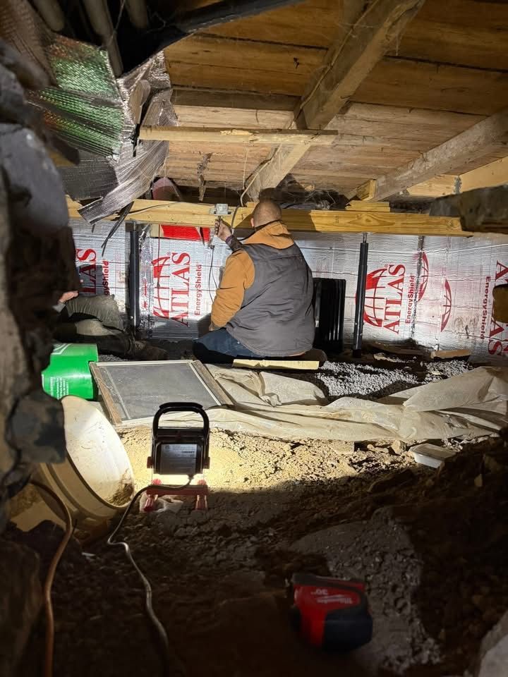 Person sits under a wooden structure; exposed insulation and tools in the crawl space.