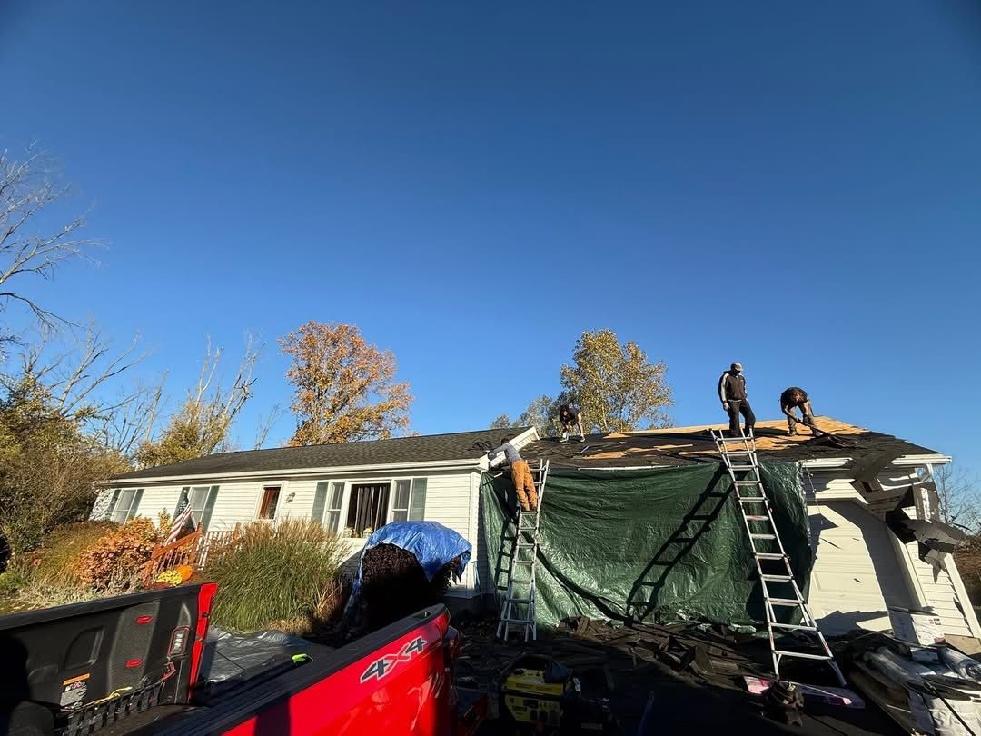 People working on a house roof under a clear blue sky. A green tarp is covering part of the structure.