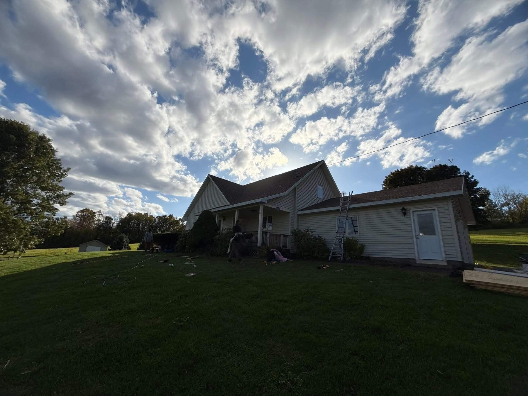 House with white siding and brown roof under a cloudy, blue sky on a grassy hill.