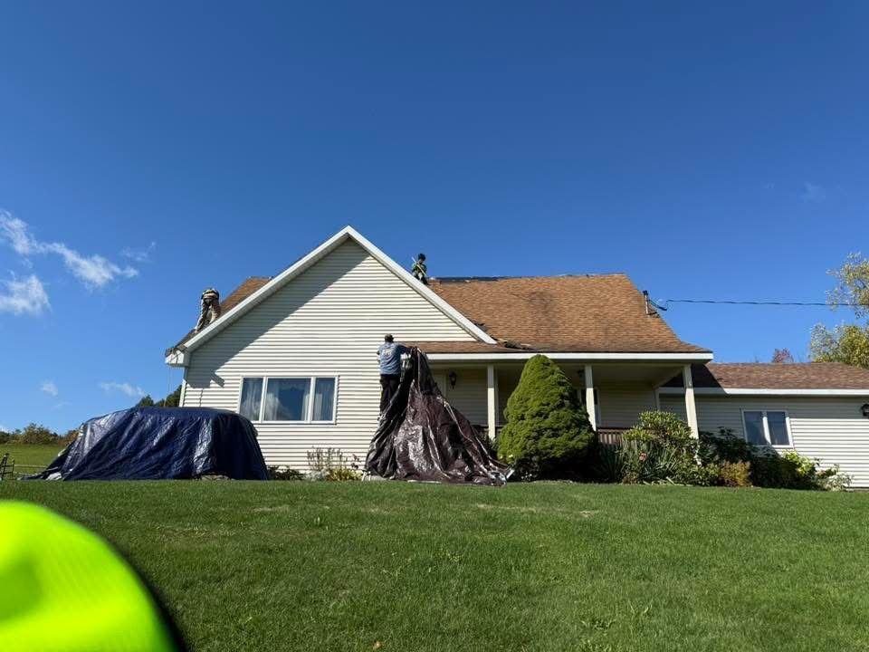 People on a roof covering it with tarps. House with green grass under a blue sky.